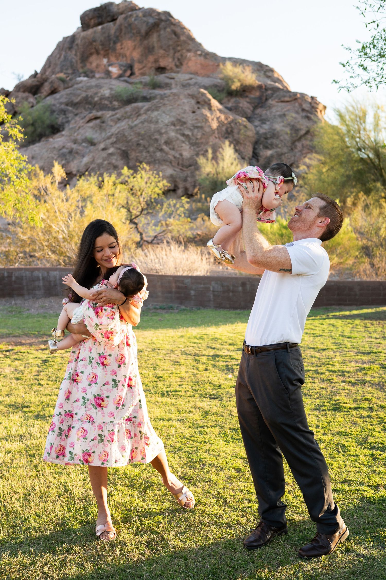 Happy family playing outdoors on green grass with rocky desert landscape, mother holding baby, father lifting toddler.