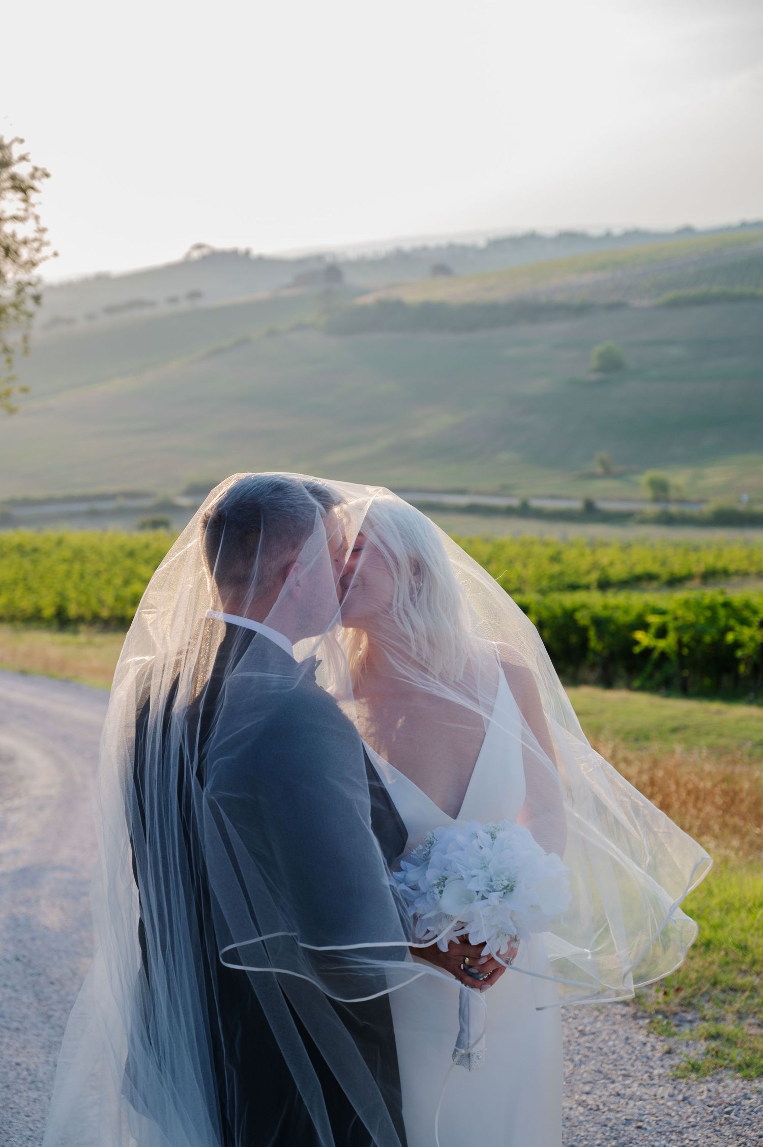 Bride and groom sharing a kiss under the bridal veil with lush green vineyard hills behind in Tuscany Italy.