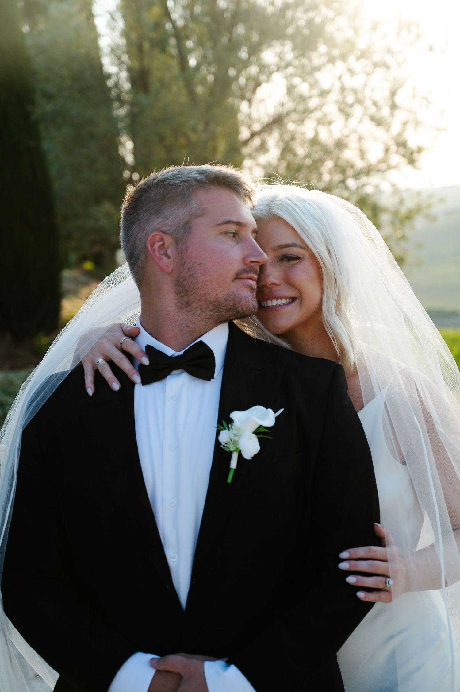 Bride in white gown and veil embraces groom in black tuxedo with bow tie and white boutonniere outdoors.