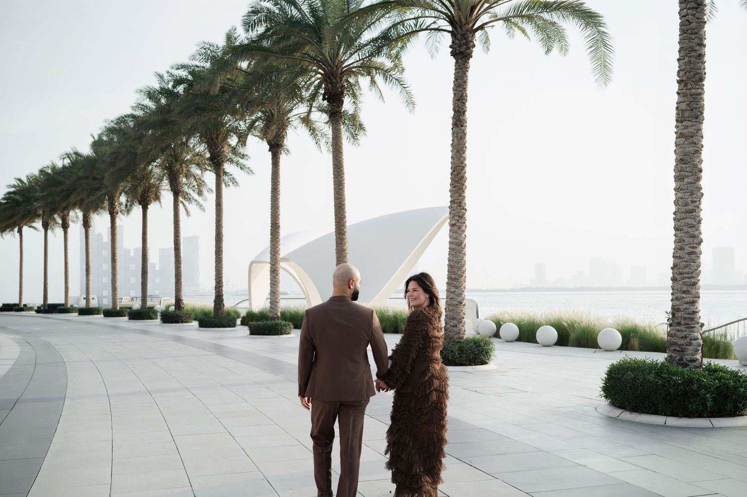 Couple walks hand-in-hand along a palm-lined waterfront promenade with a modern cityscape in the background.