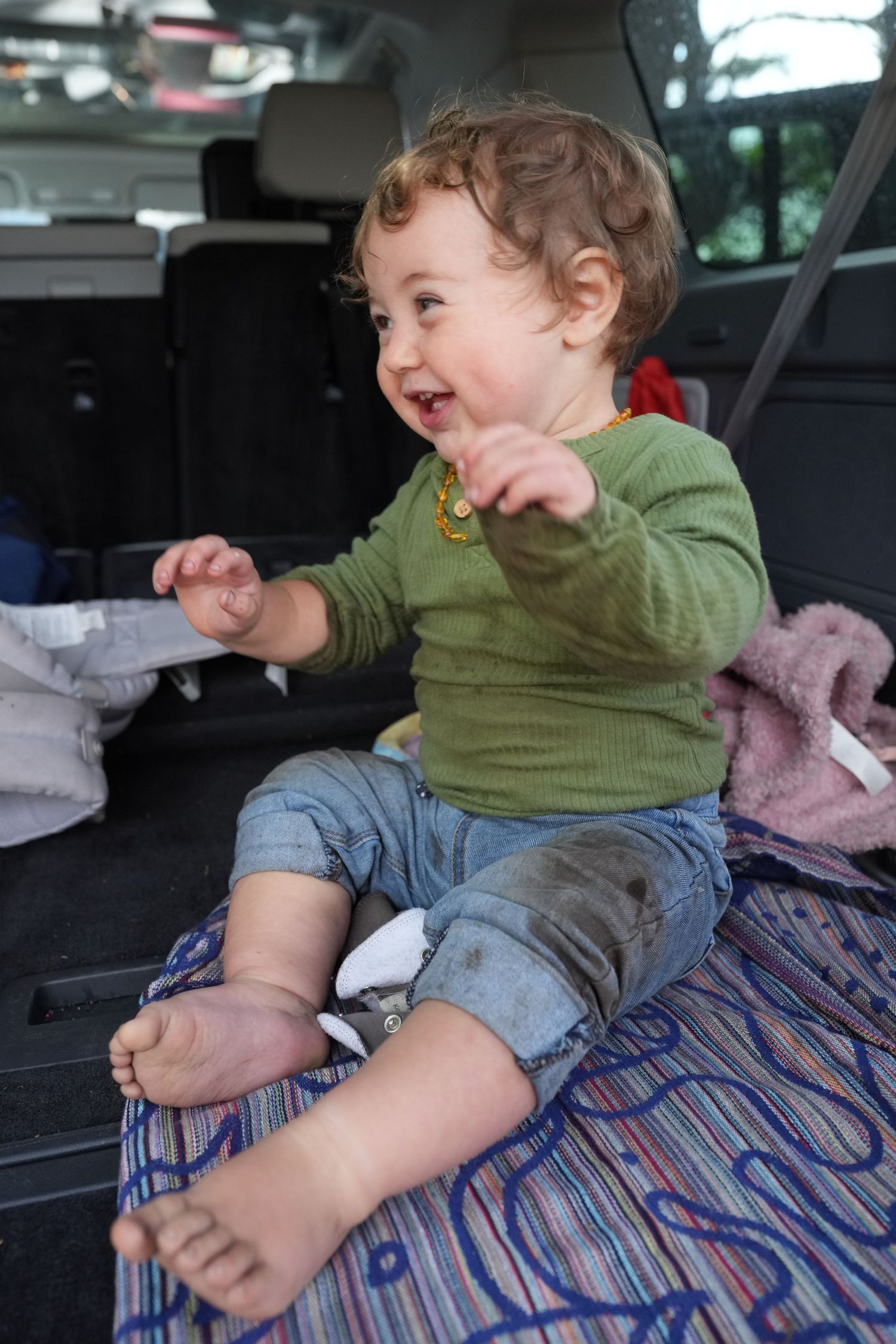 Happy toddler in green shirt sitting in car trunk with dirty feet laughing.