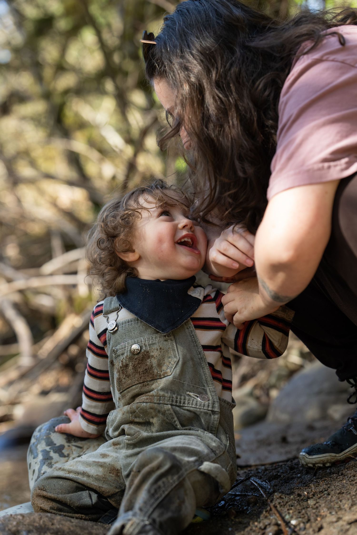 Mother bends down smiling at laughing toddler in dirty overalls near a woodland stream.