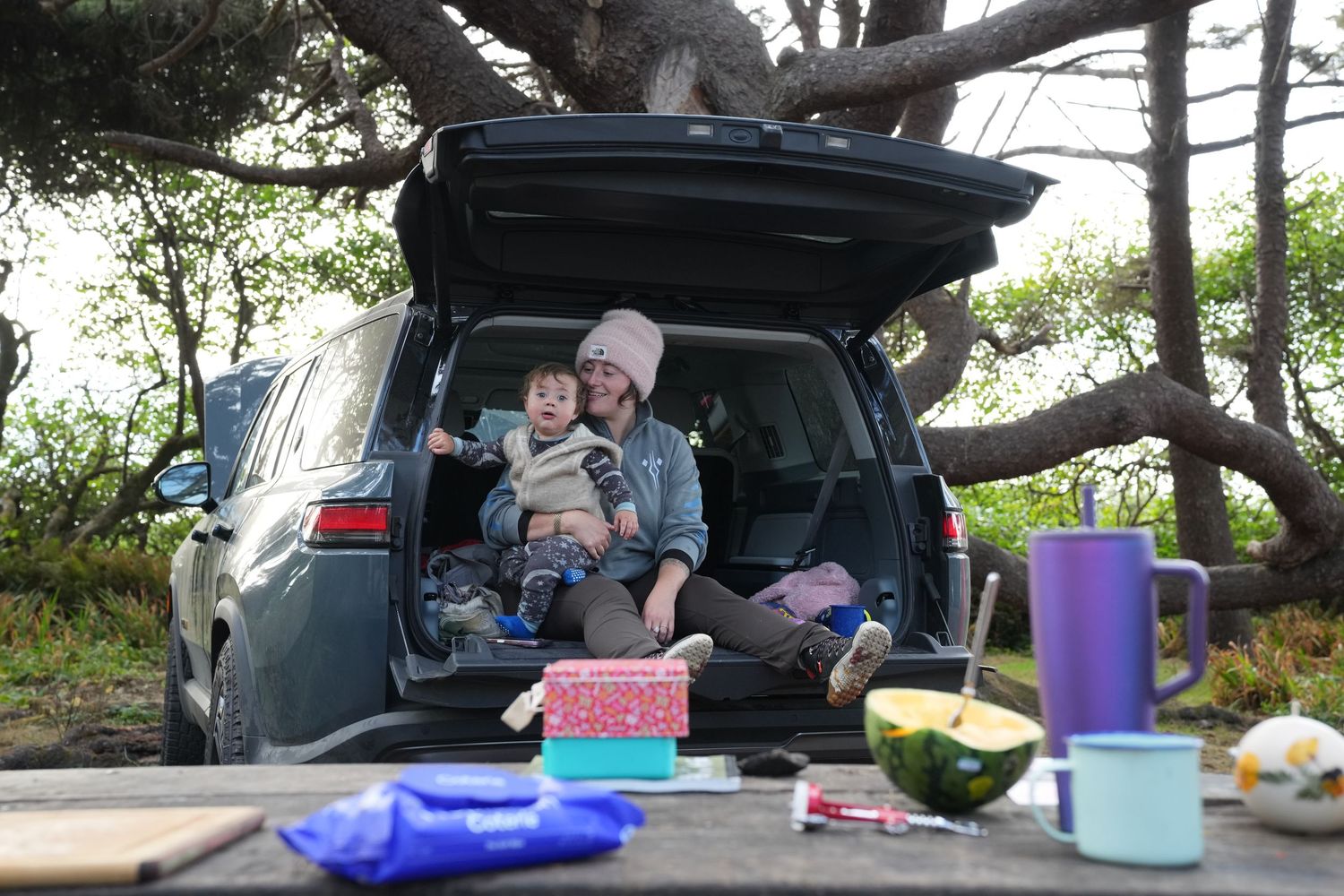 Woman in pink beanie sitting in car trunk with toddler at outdoor campsite.