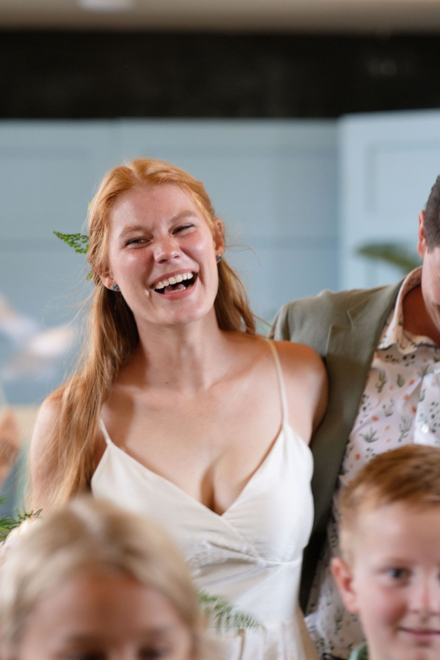 Radiant redhead bride in white slip dress laughing joyfully with fern hair accessory at wedding reception.