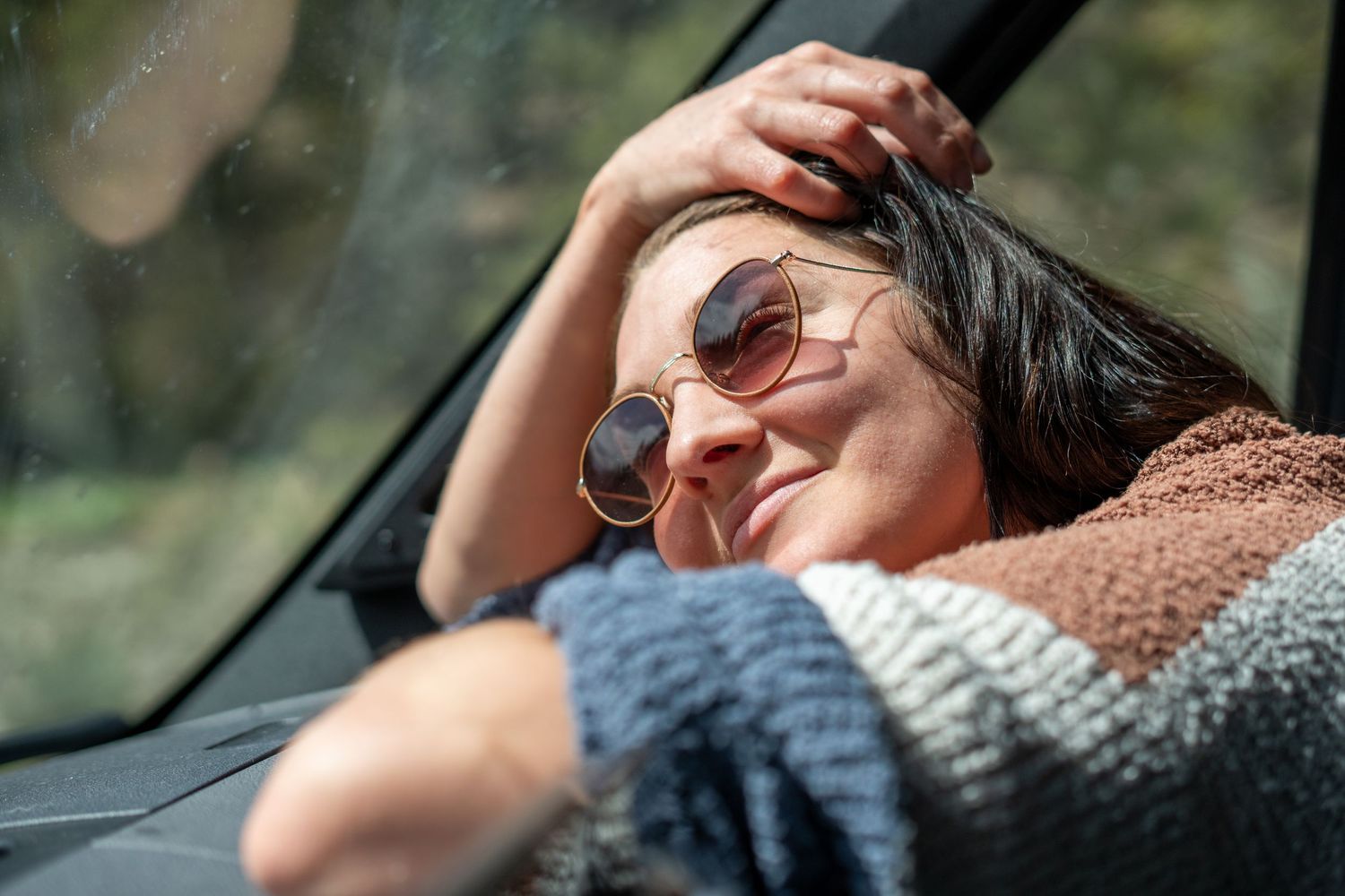 Relaxed woman in sunglasses leaning forward on a car dashboard enjoying a sunny road trip outdoors.