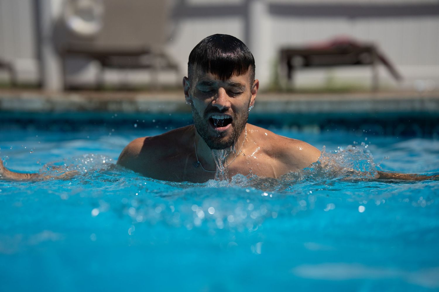 Young man emerging from a bright blue swimming pool with water dripping from his face.