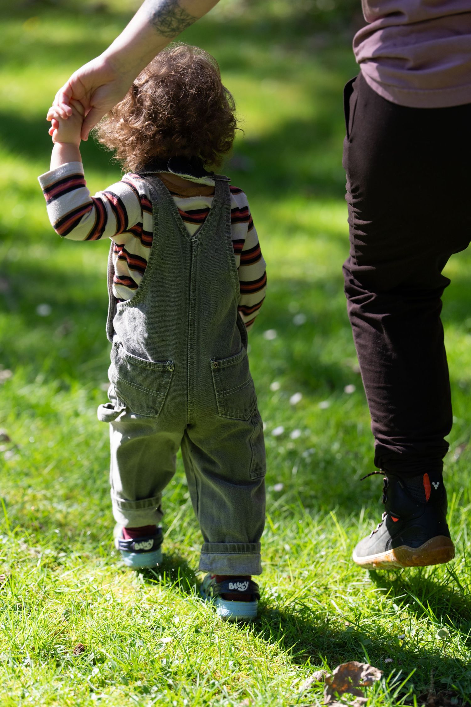 Toddler in green overalls holds adult hand while walking on lush grass in sunny park.
