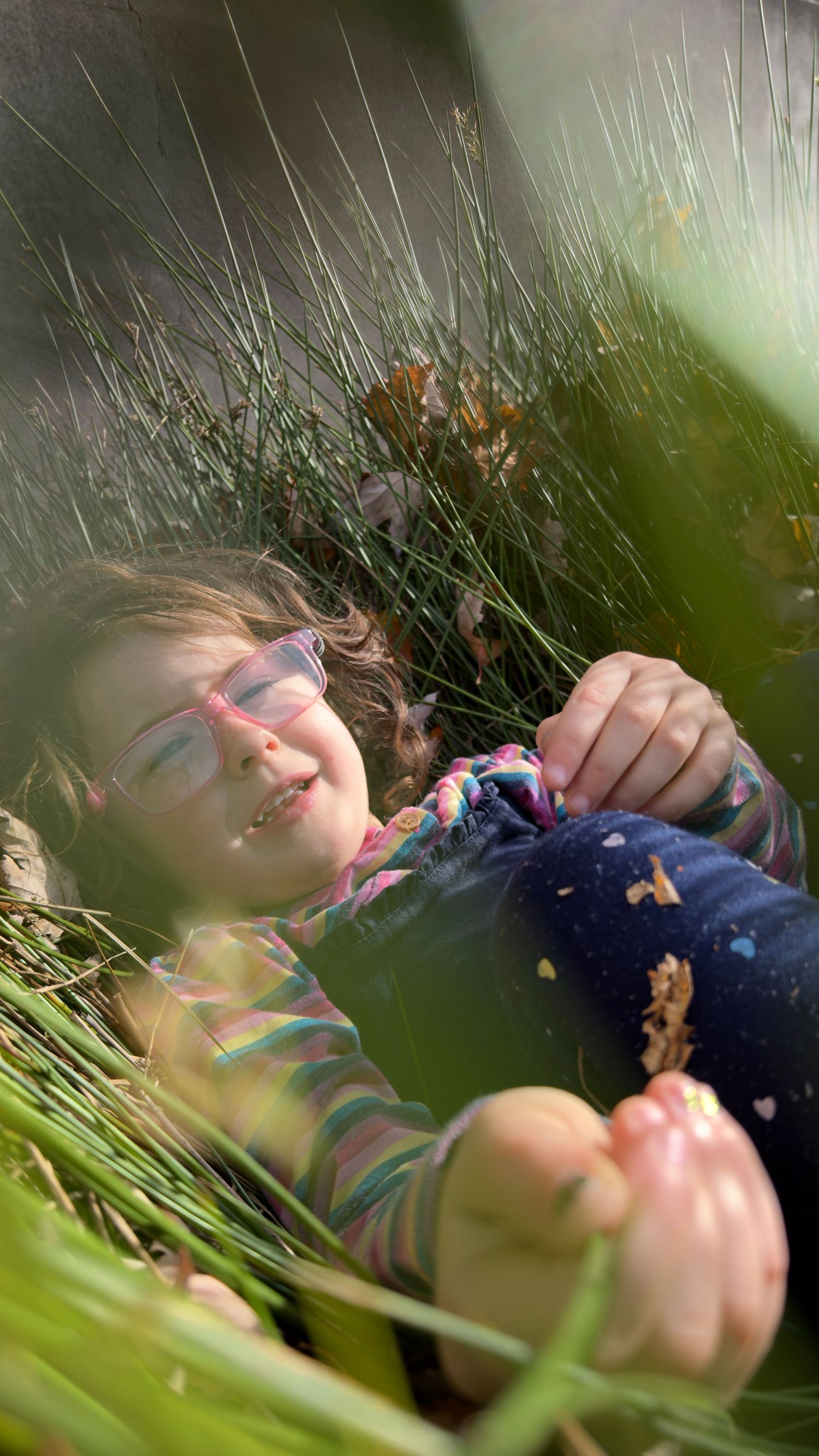 Young girl with pink glasses lying peacefully among green grass and autumn leaves in soft natural sunlight.