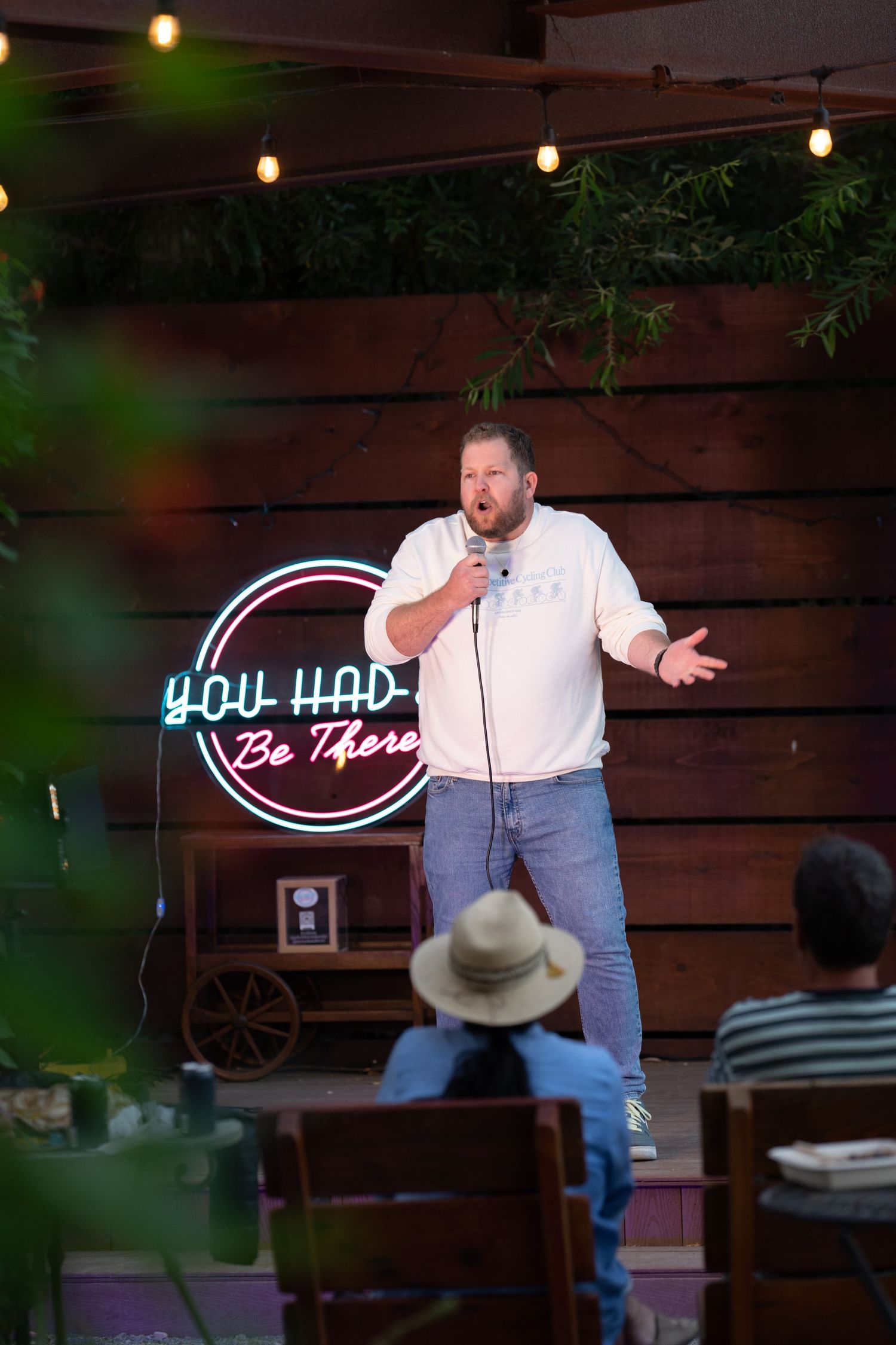 Male comedian in white shirt performing on stage with a neon You Had To Be There sign at outdoor venue.