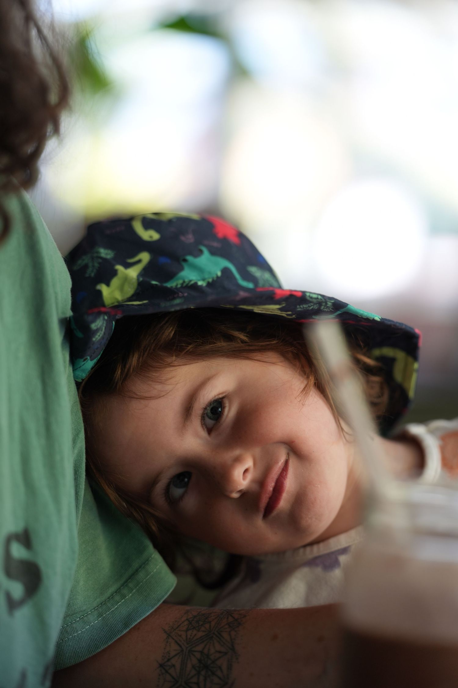Cute toddler in a colorful dinosaur bucket hat smiling while leaning against an adult.