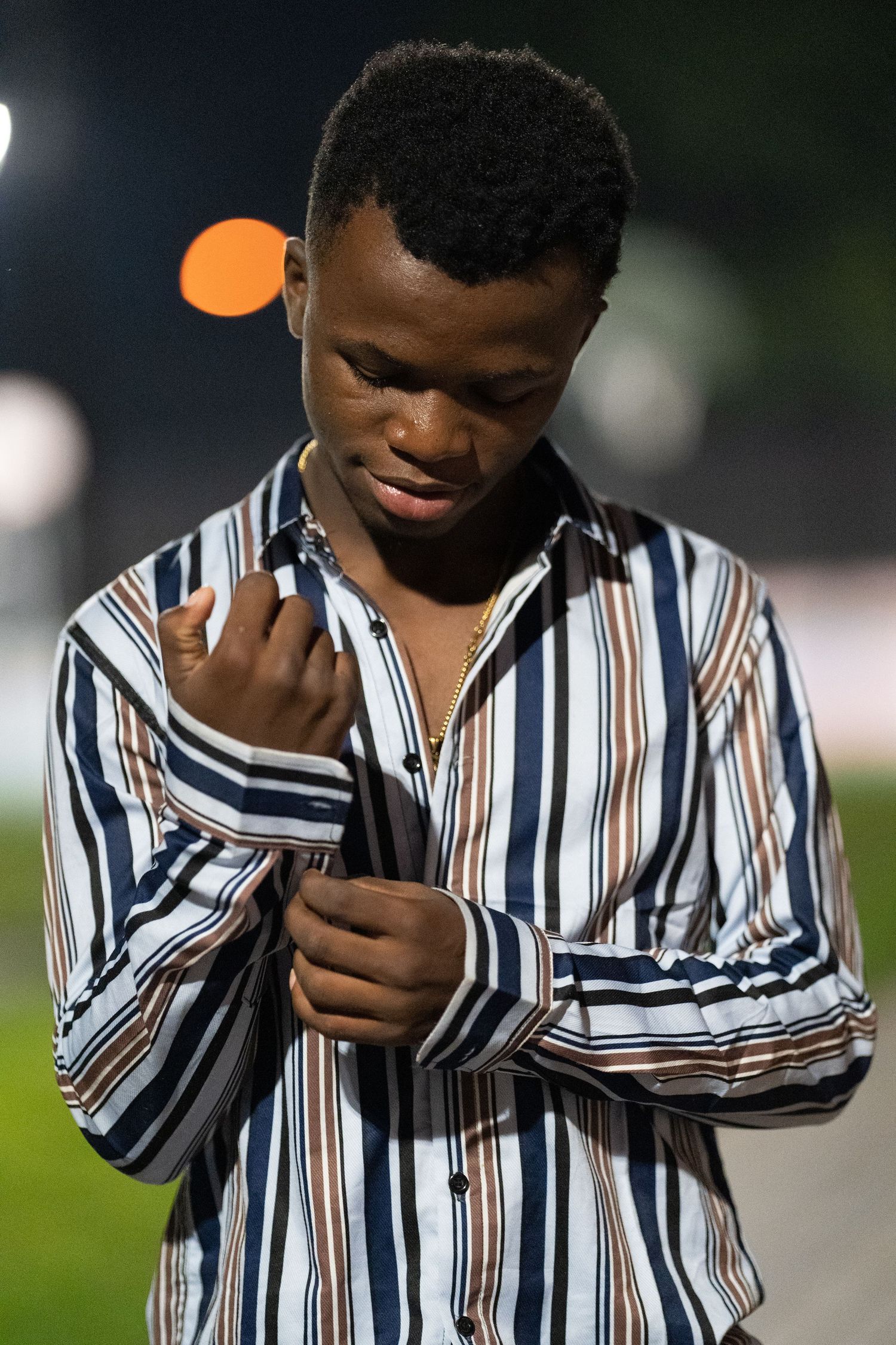 Young black man in striped shirt adjusting cuffs at night with bokeh lights in the background.