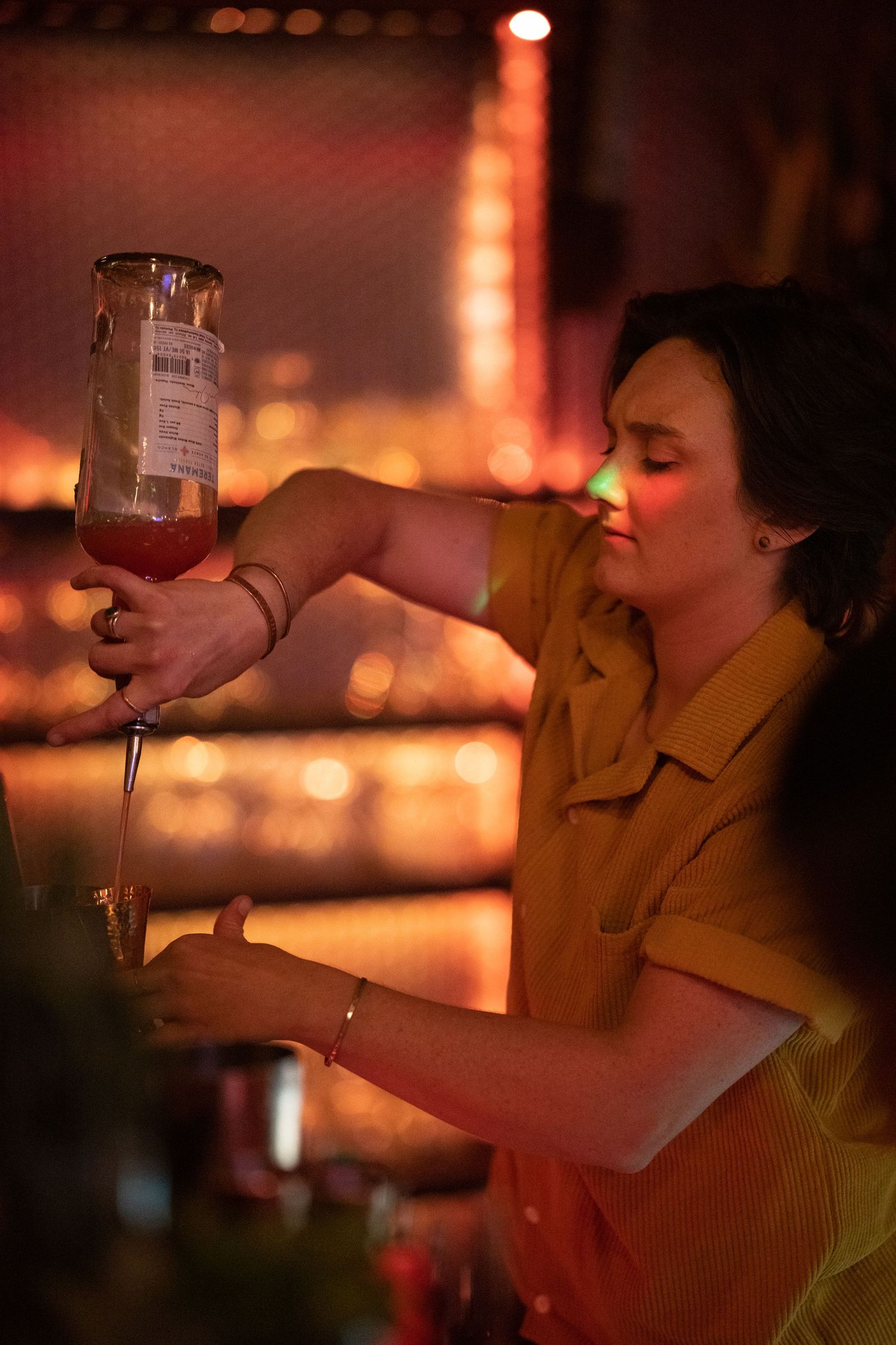 Female bartender in yellow button up shirt pouring red cocktail from shaker at a dimly lit bar.