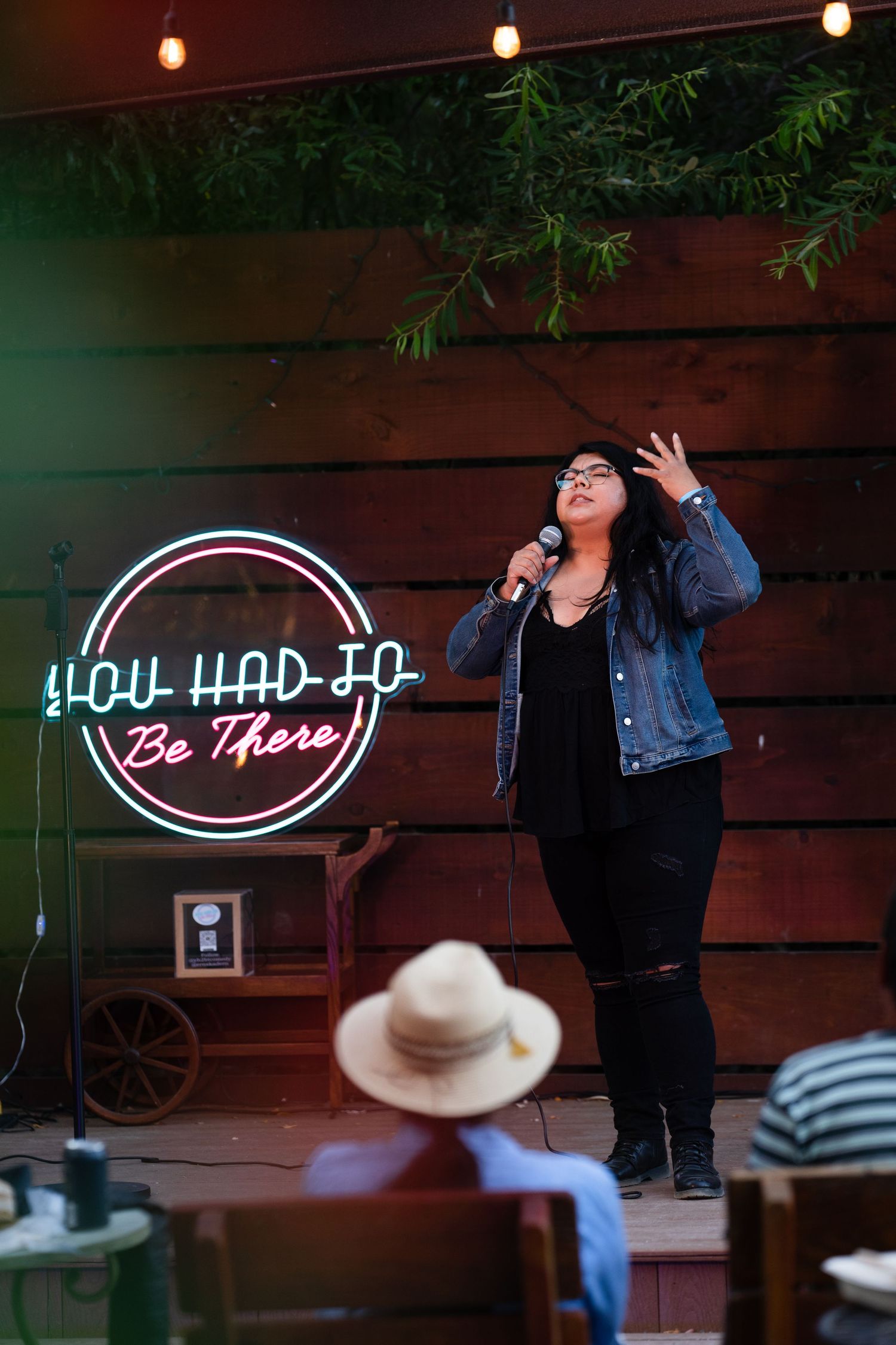Female comedian performing on stage with a neon You Had To Be There sign at an outdoor comedy show.