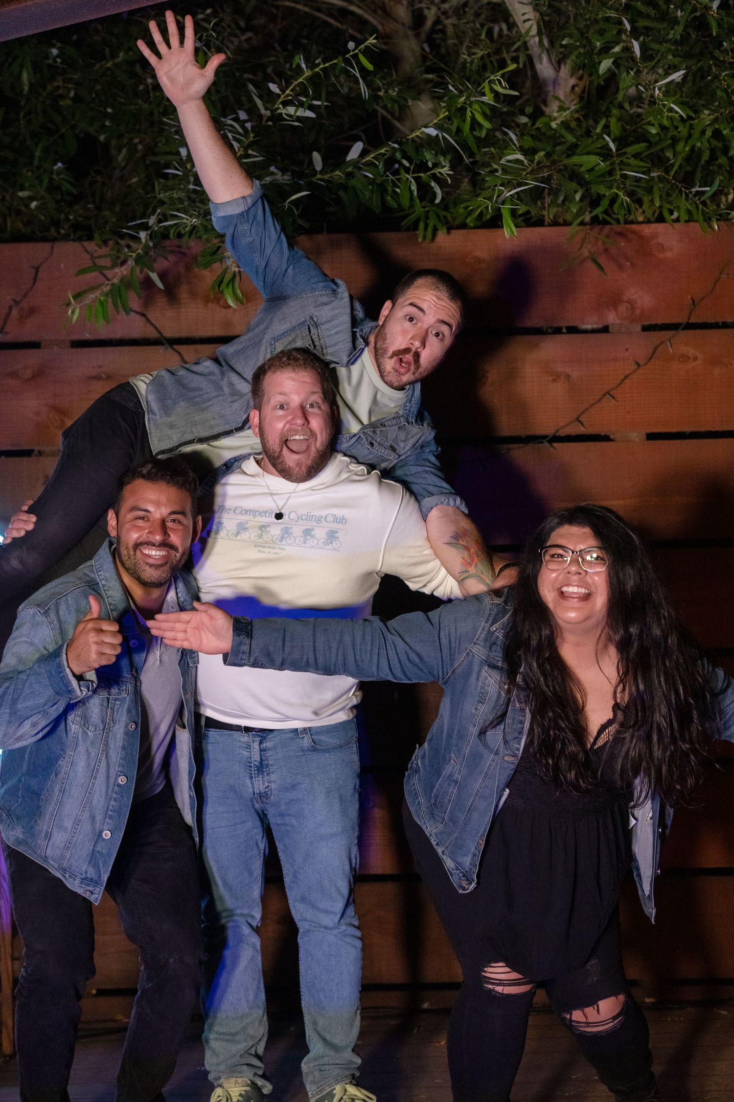 Four energetic comedians and performers celebrating and posing playfully in front of a wooden fence at night.