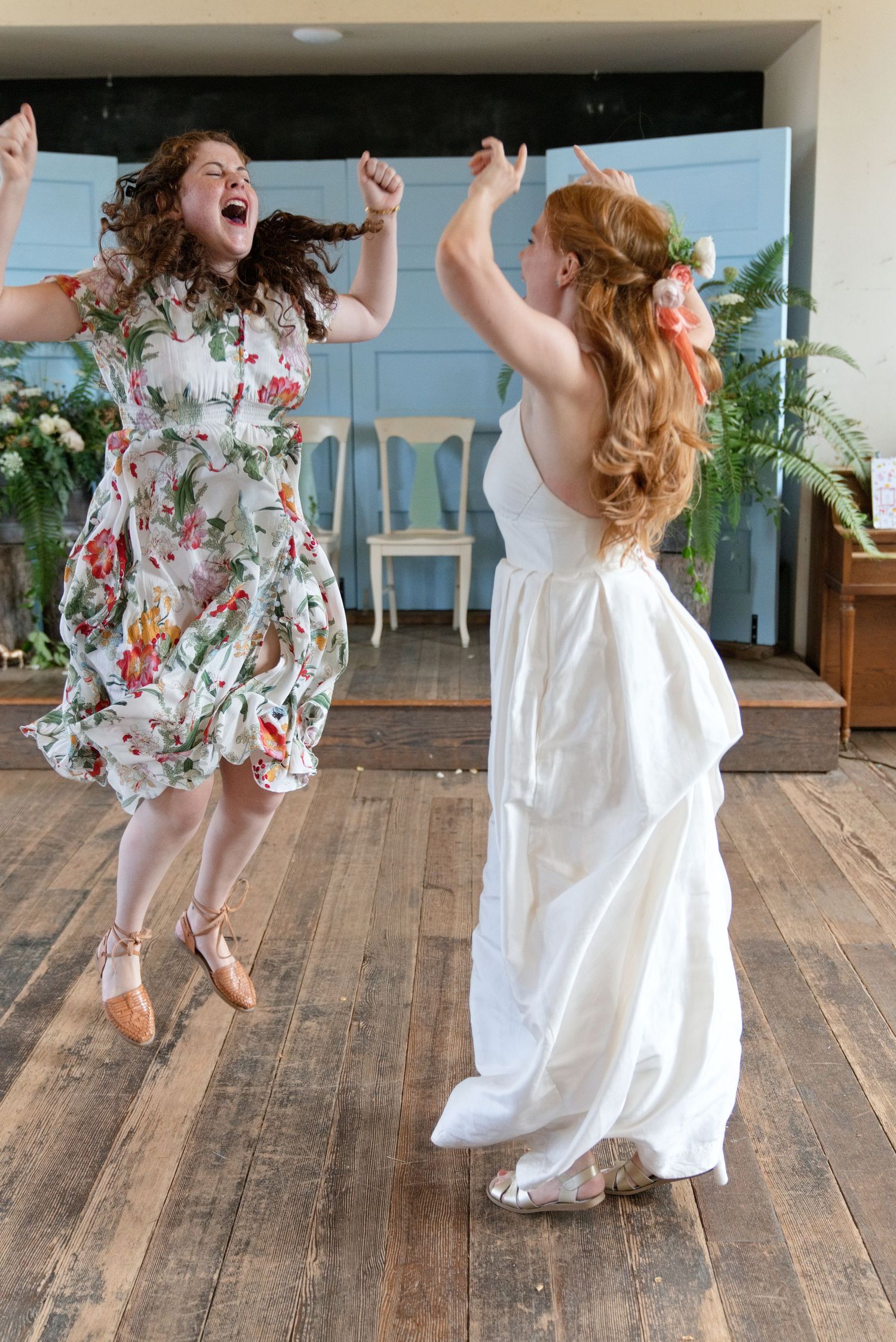 Joyful bride in white gown dancing with bridesmaid in floral dress on wooden dance floor.