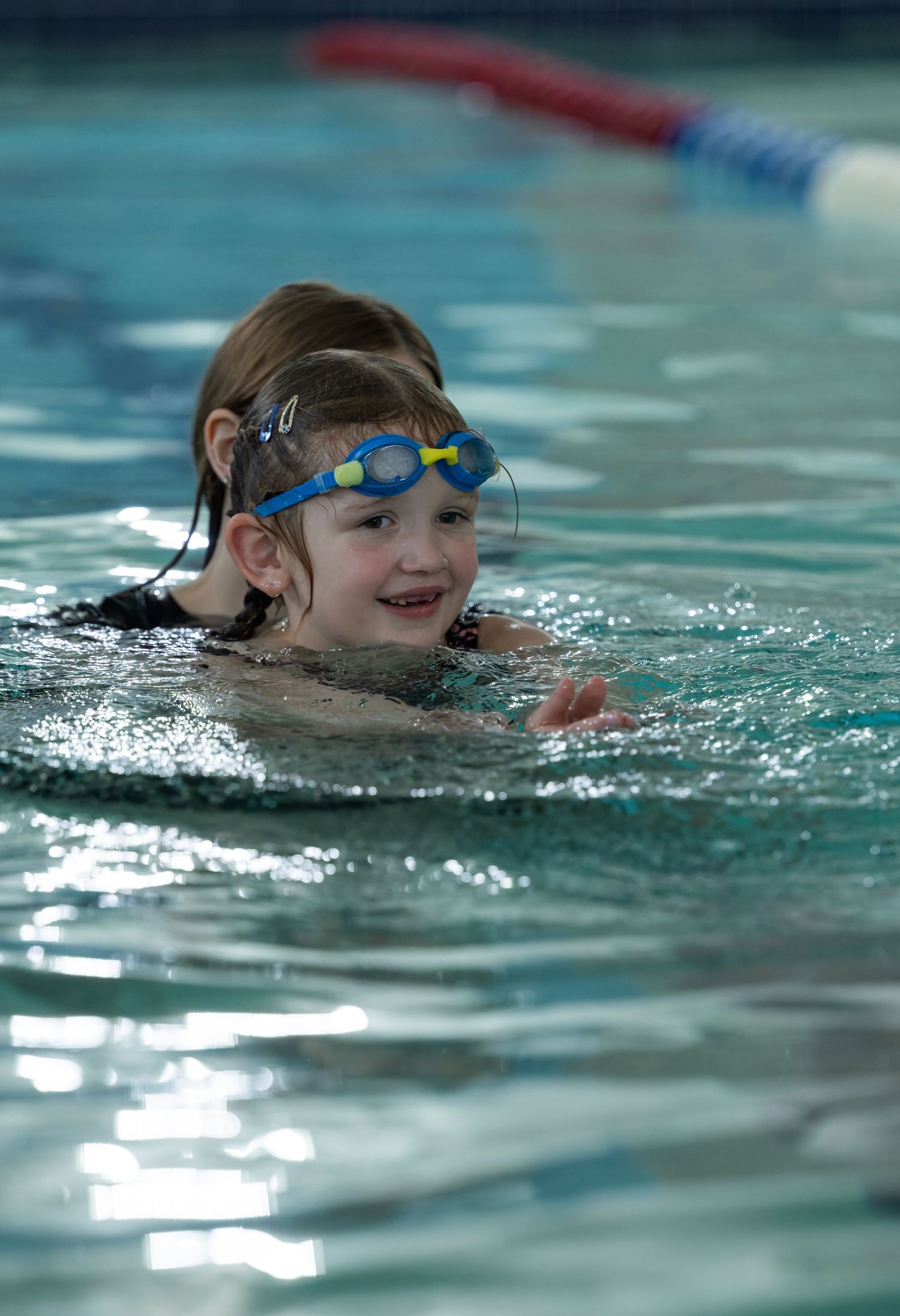 Happy young girl wearing blue swim goggles swimming in an indoor pool smiling during a swim lesson.