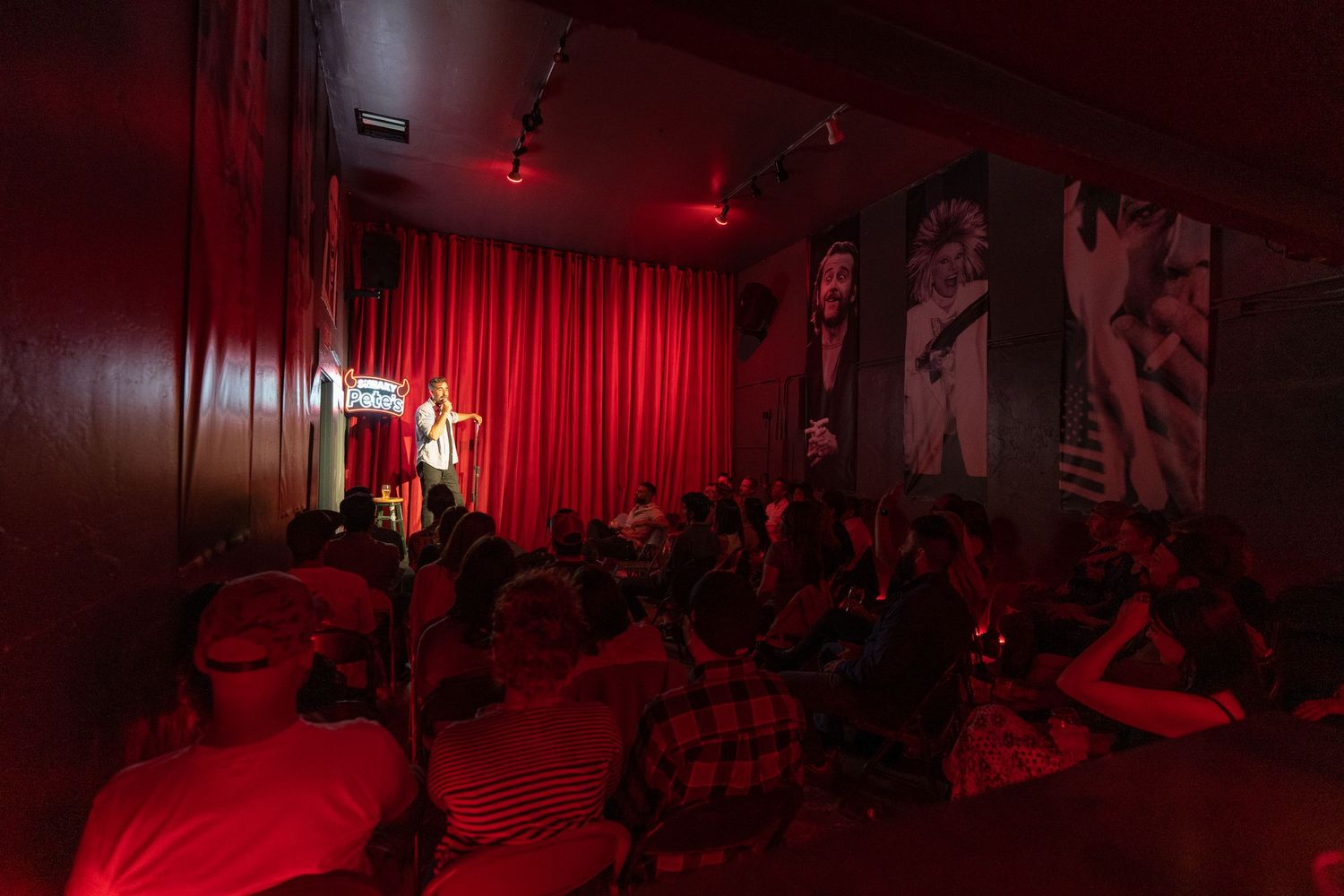 Audience watching a performer on stage in a red-lit intimate comedy club venue.