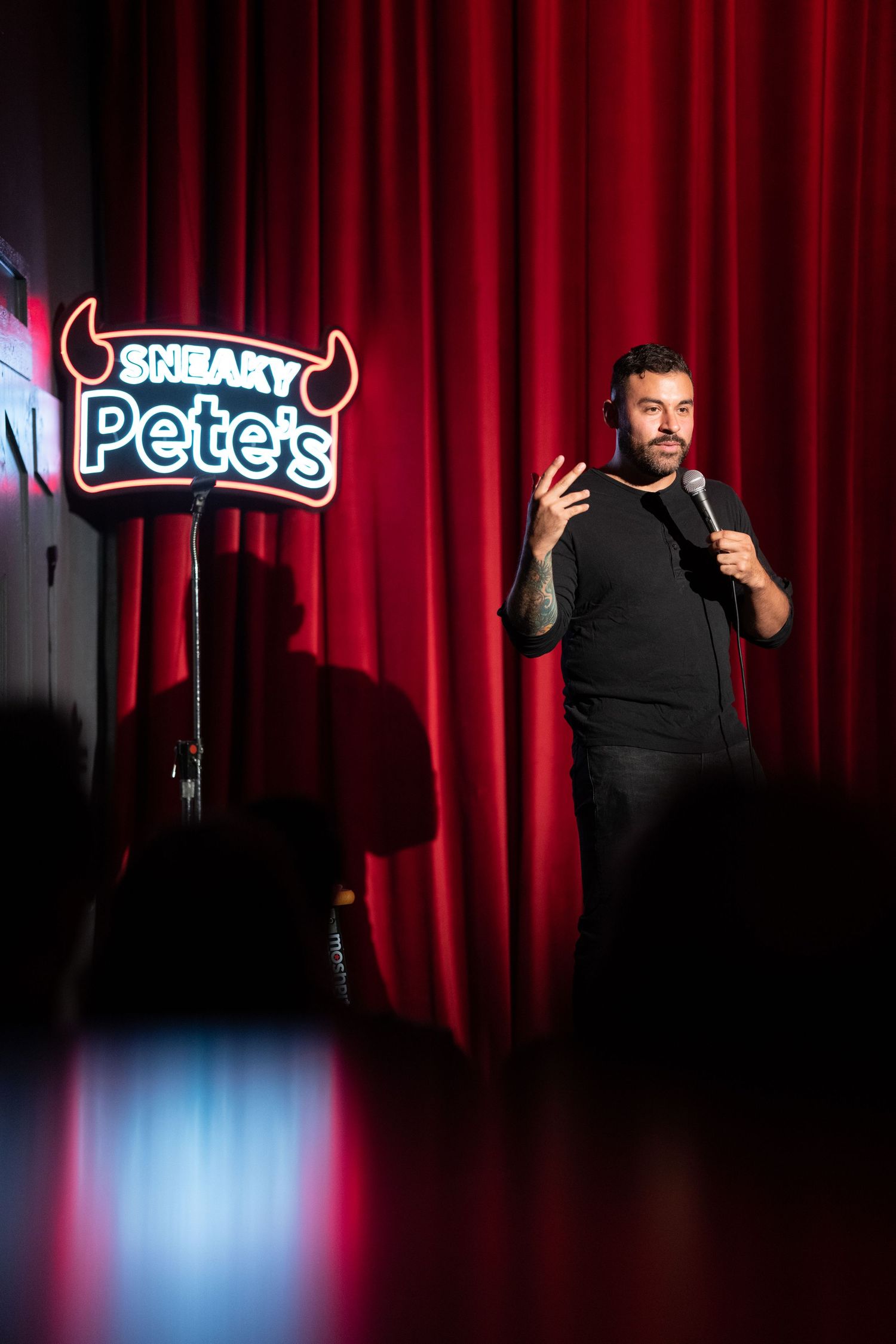Male stand-up comedian performing on stage at Sneaky Pete's comedy club with red curtain backdrop.