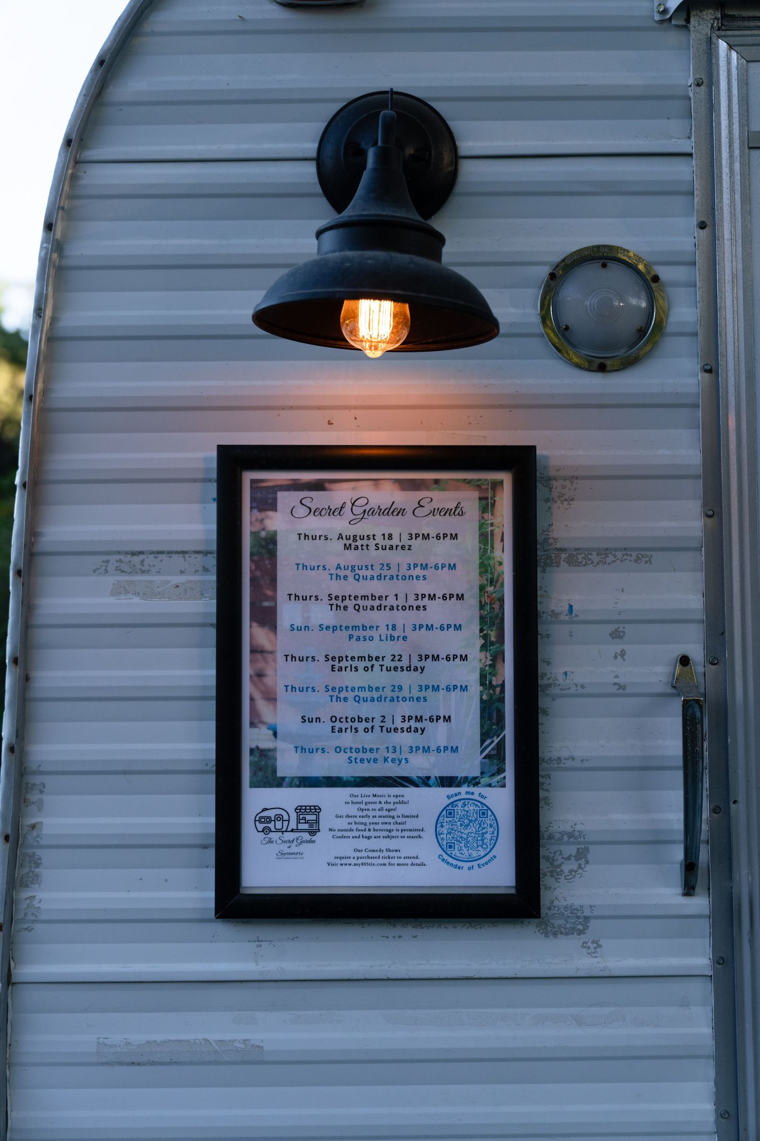 Illuminated vintage trailer exterior with a framed Secret Garden Events schedule sign and Edison bulb wall sconce at dusk.