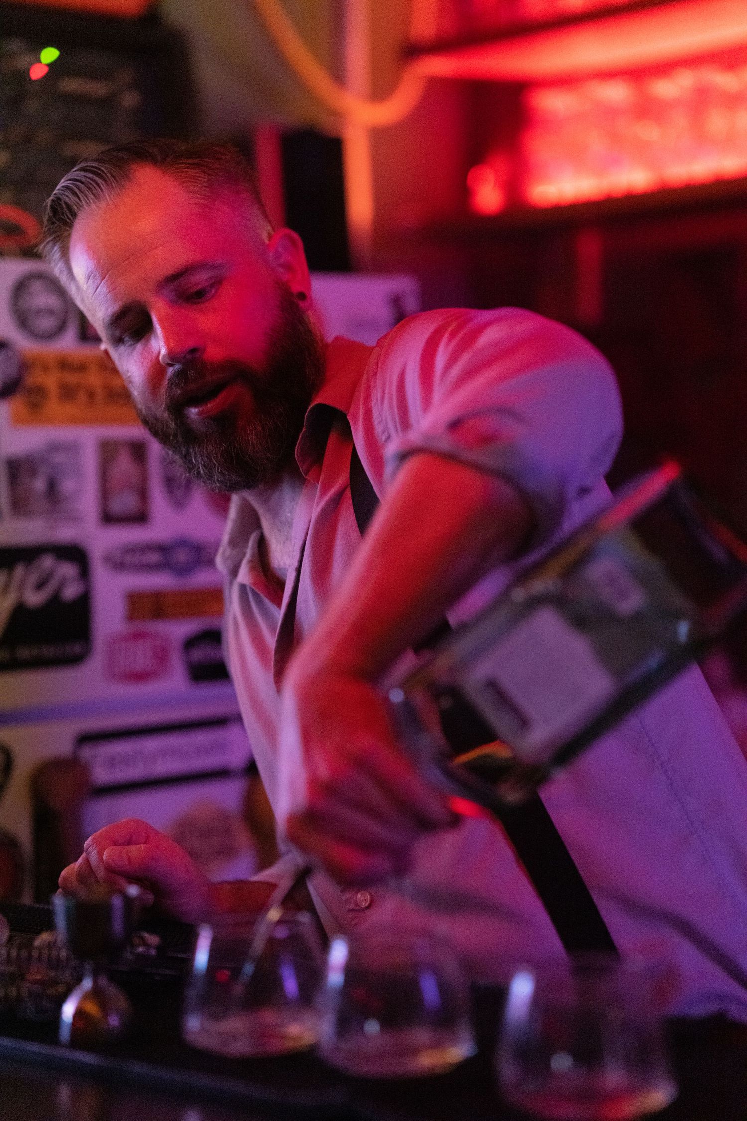 Bearded male bartender preparing cocktails at a neon-lit bar with colorful drink glasses.