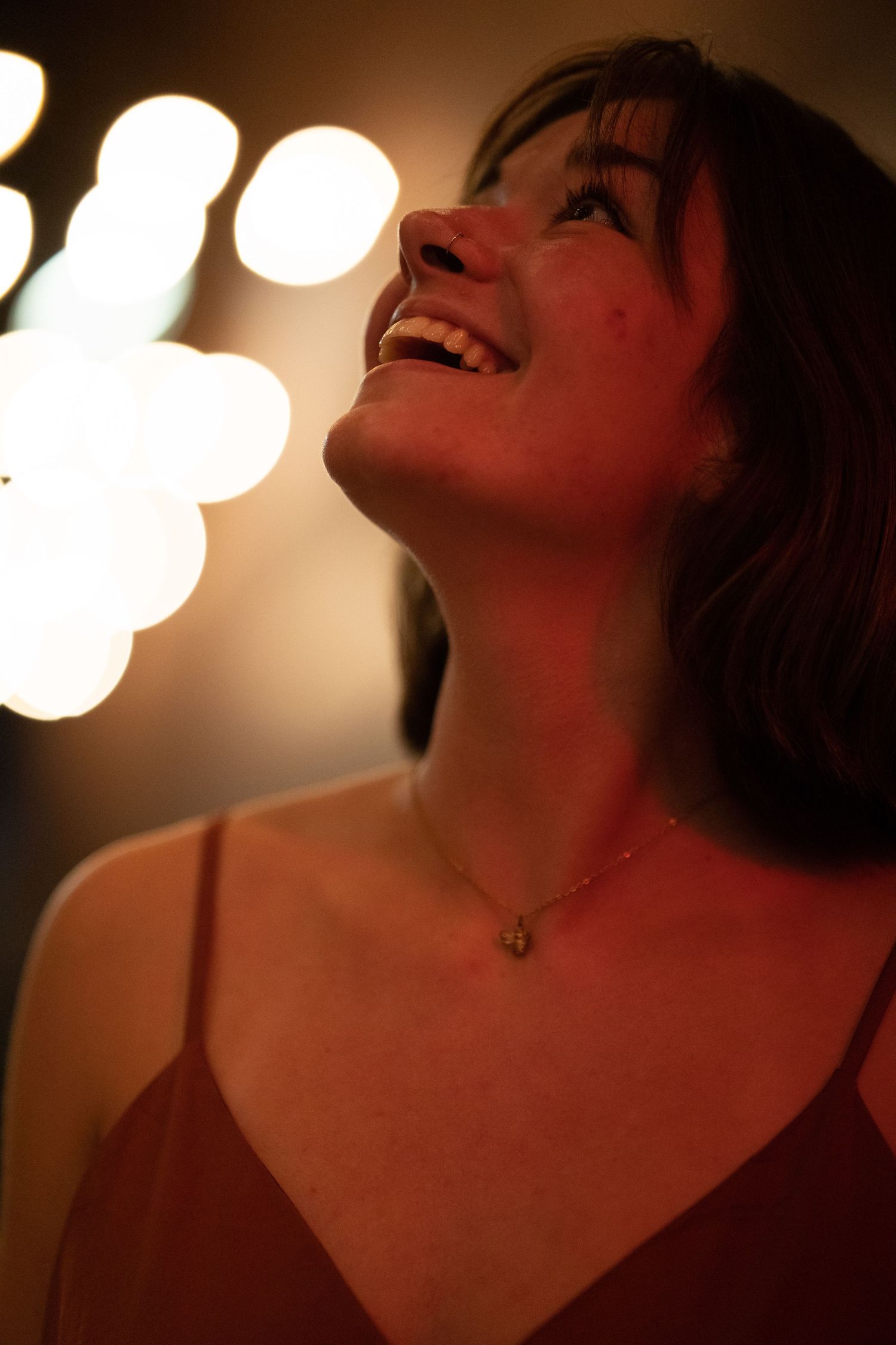 Young woman in red dress laughs joyfully looking up at warm bokeh lights in background.
