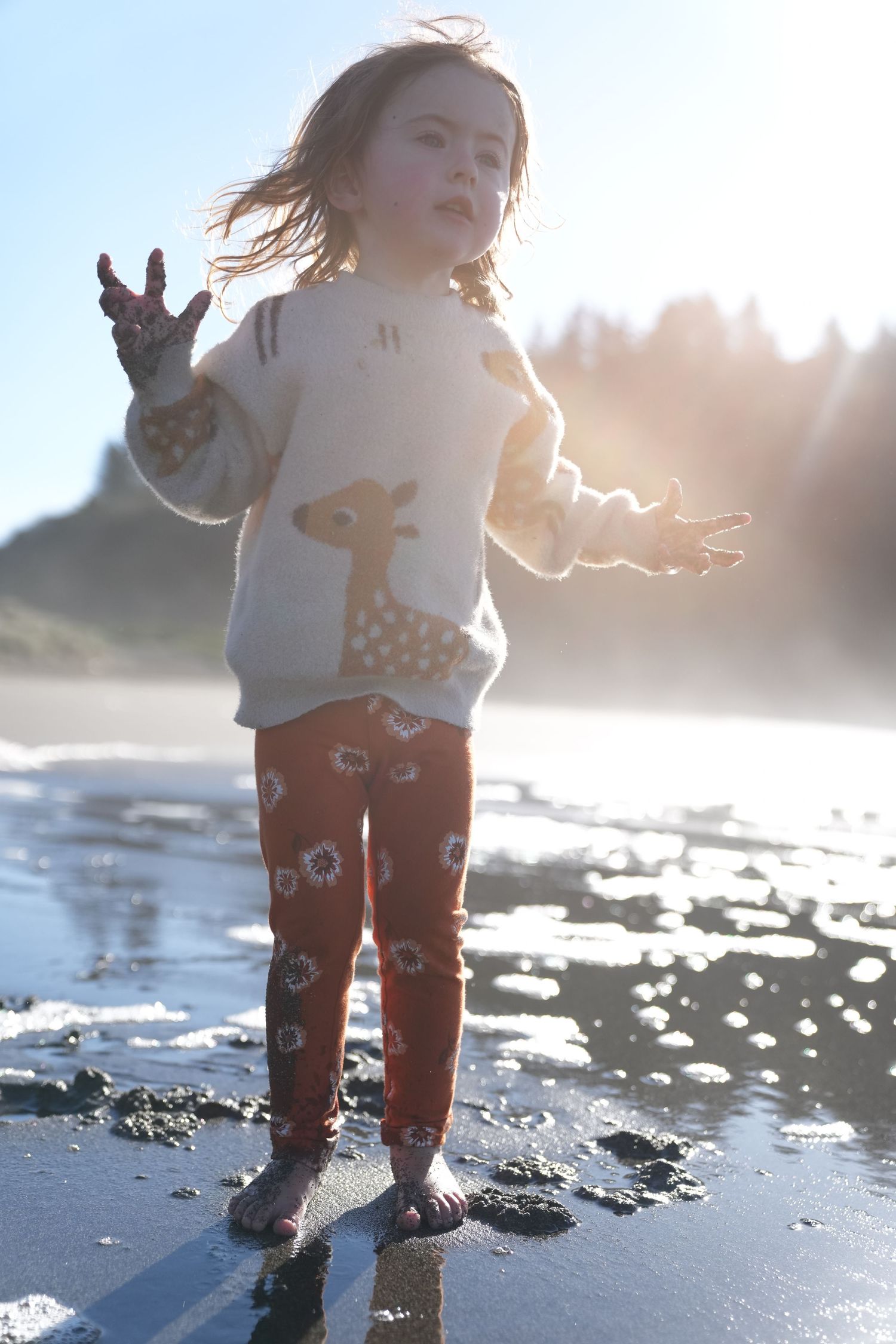 Young girl in giraffe sweater and red pants standing on a sunny beach with arms raised.