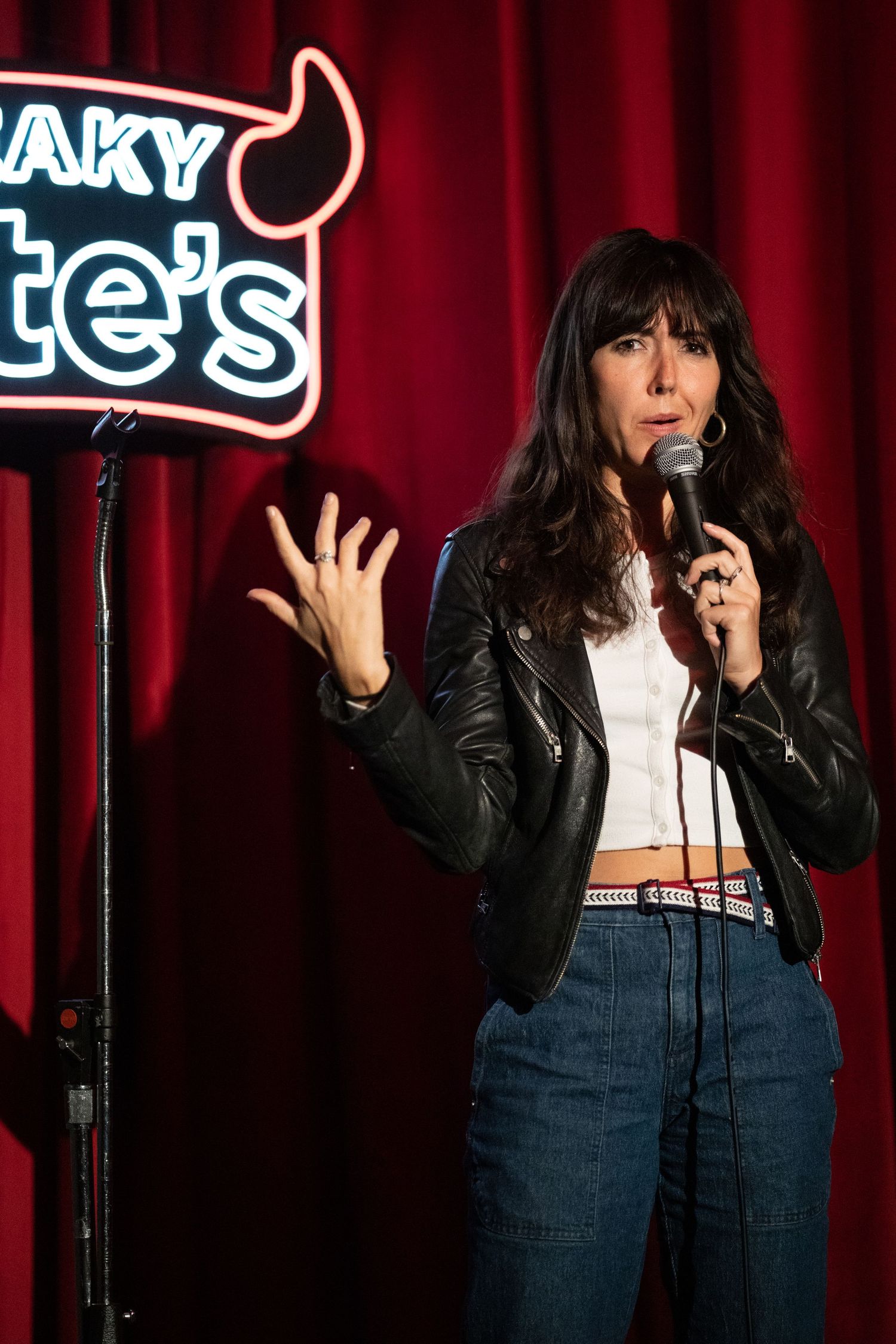 Female comedian performing stand-up on stage at Sneaky Pete's wearing leather jacket and jeans.