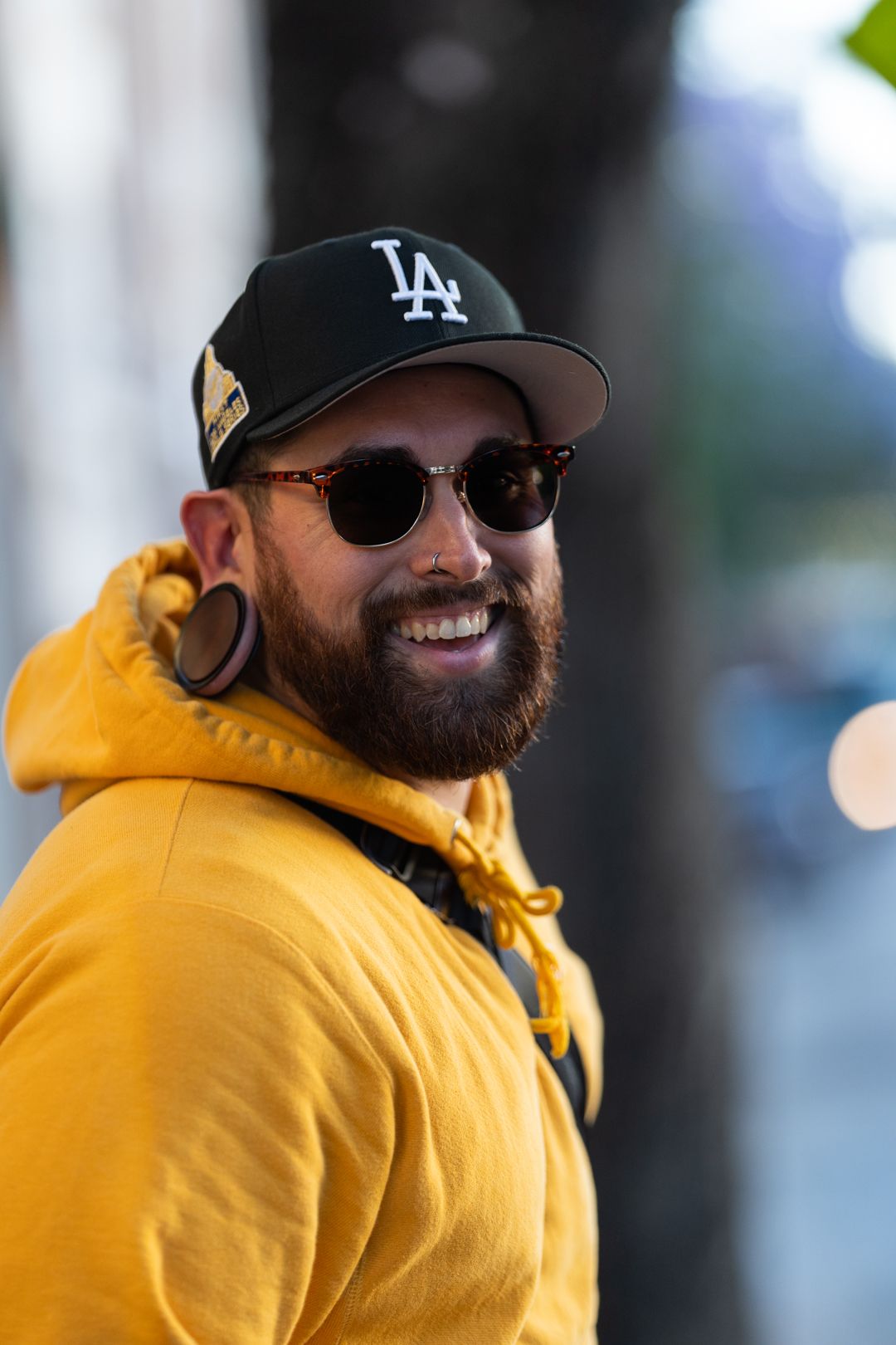 Smiling bearded man wearing LA Dodgers cap, sunglasses, and yellow hoodie outdoors.