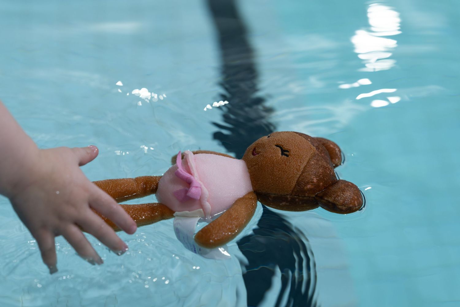 Small teddy bear in a pink swimsuit floating in a swimming pool near a child's hand.