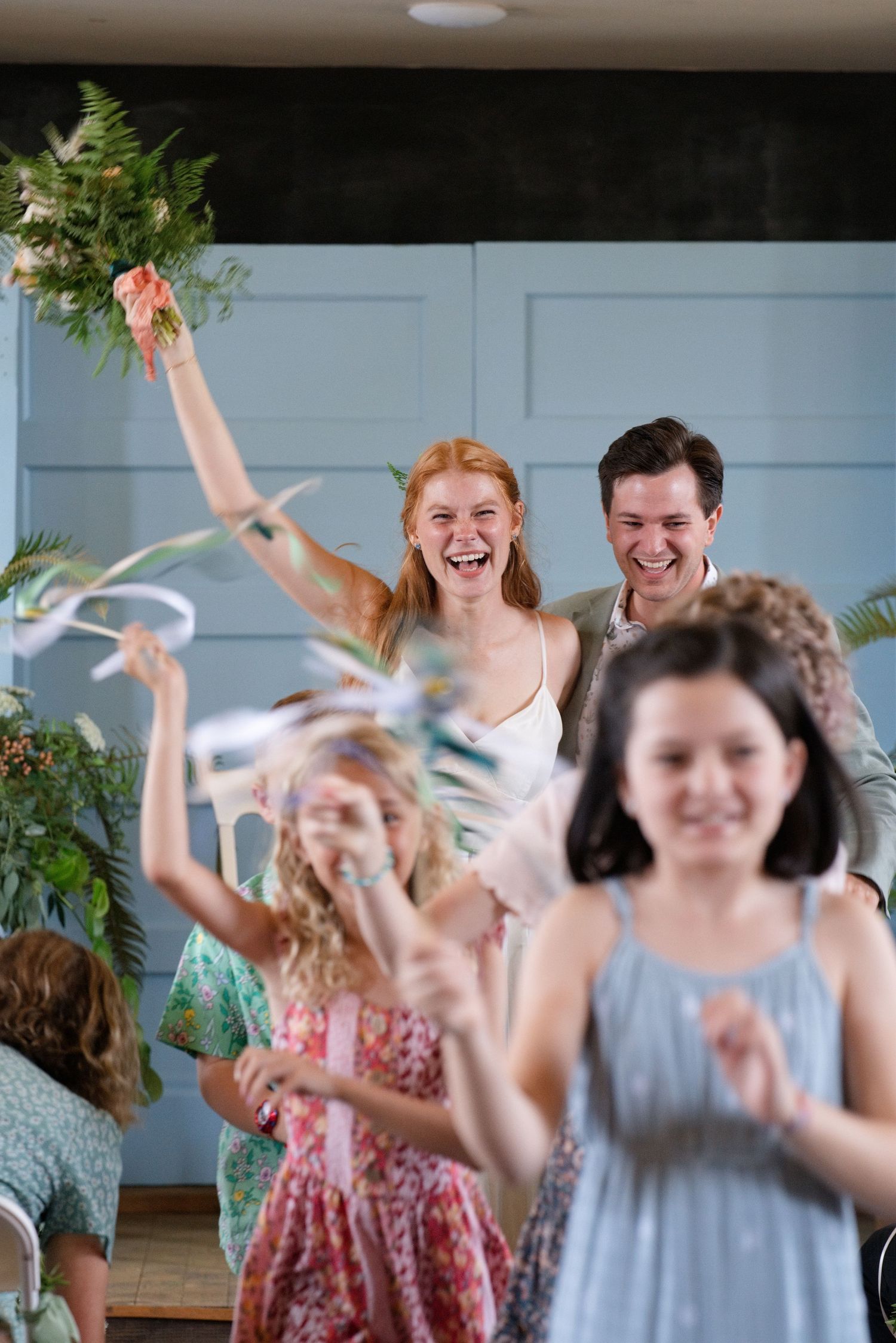 Joyful bride raising bouquet while laughing with groom during festive wedding reception celebration indoors.