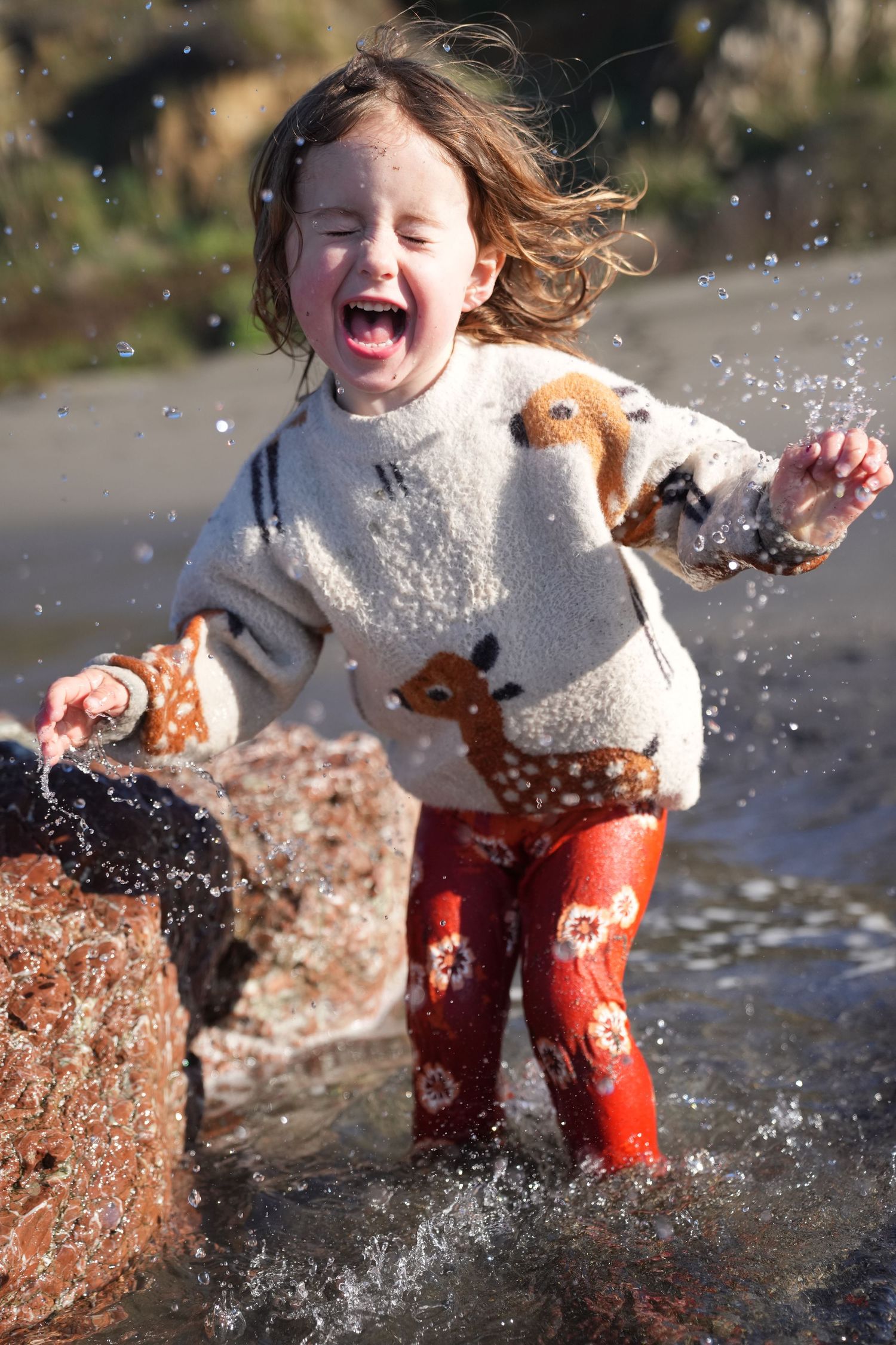 Excited young girl in animal sweater and red boots laughing while splashing in beach water.