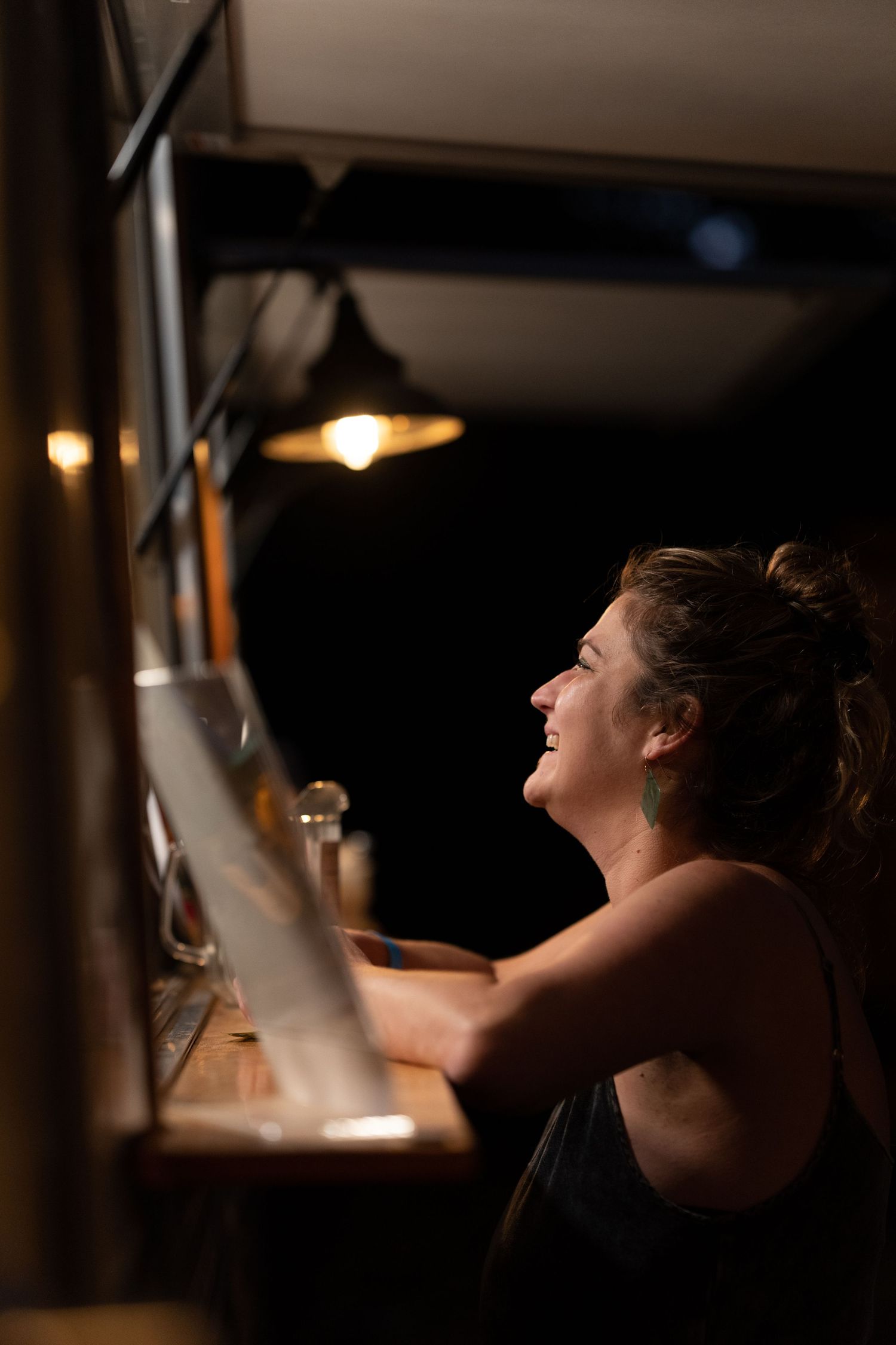Woman laughing joyfully at a dimly lit bar counter illuminated by warm pendant lights at night.