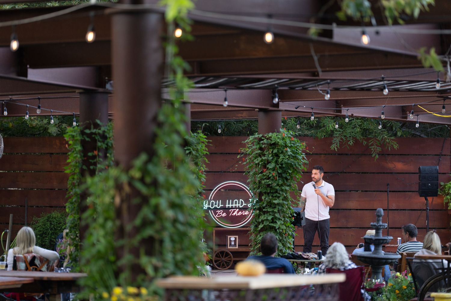 Comedian performing on an outdoor stage at a garden venue with string lights and lush greenery at night.