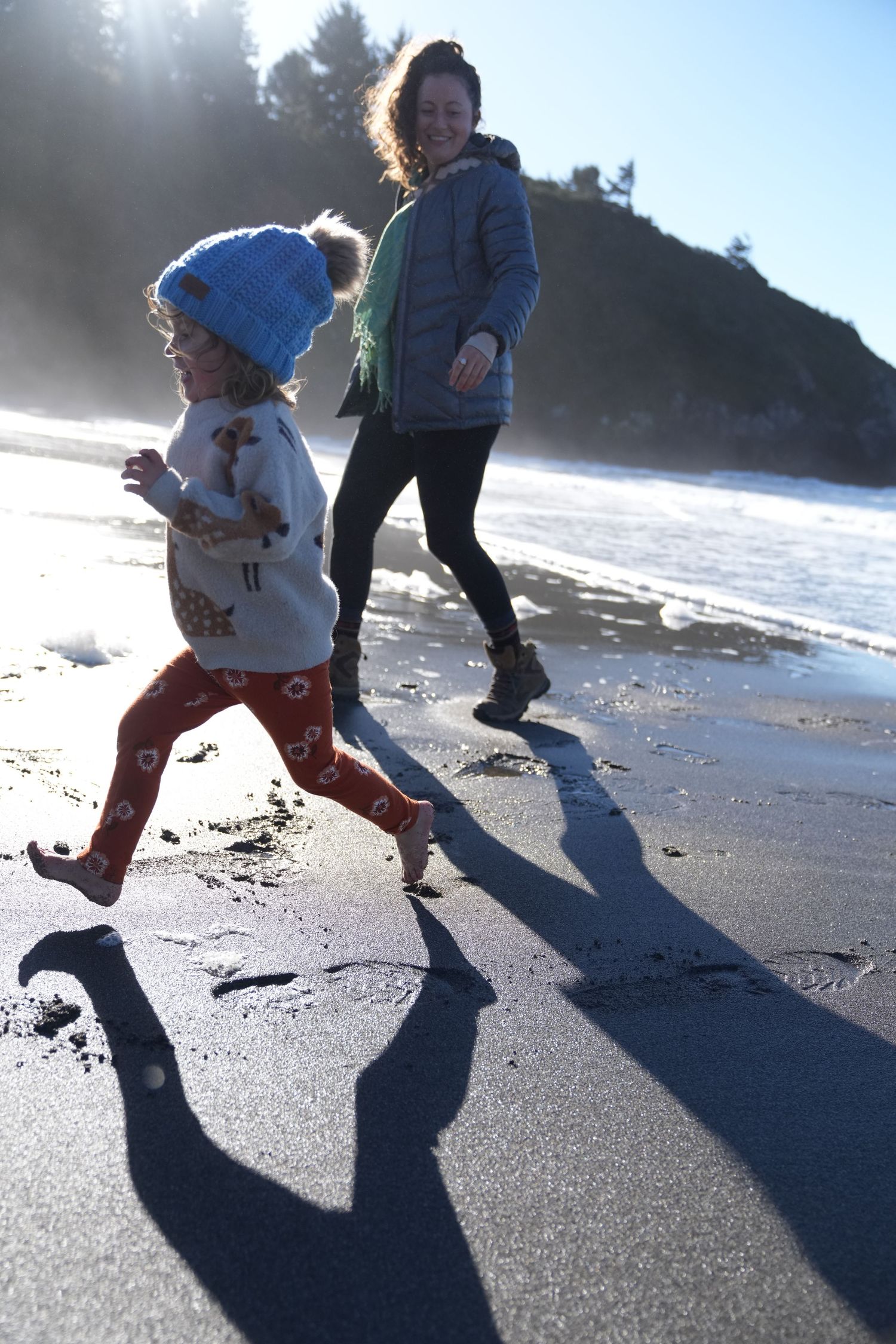 Mother and young child running barefoot on a sandy beach in bright winter sunlight.