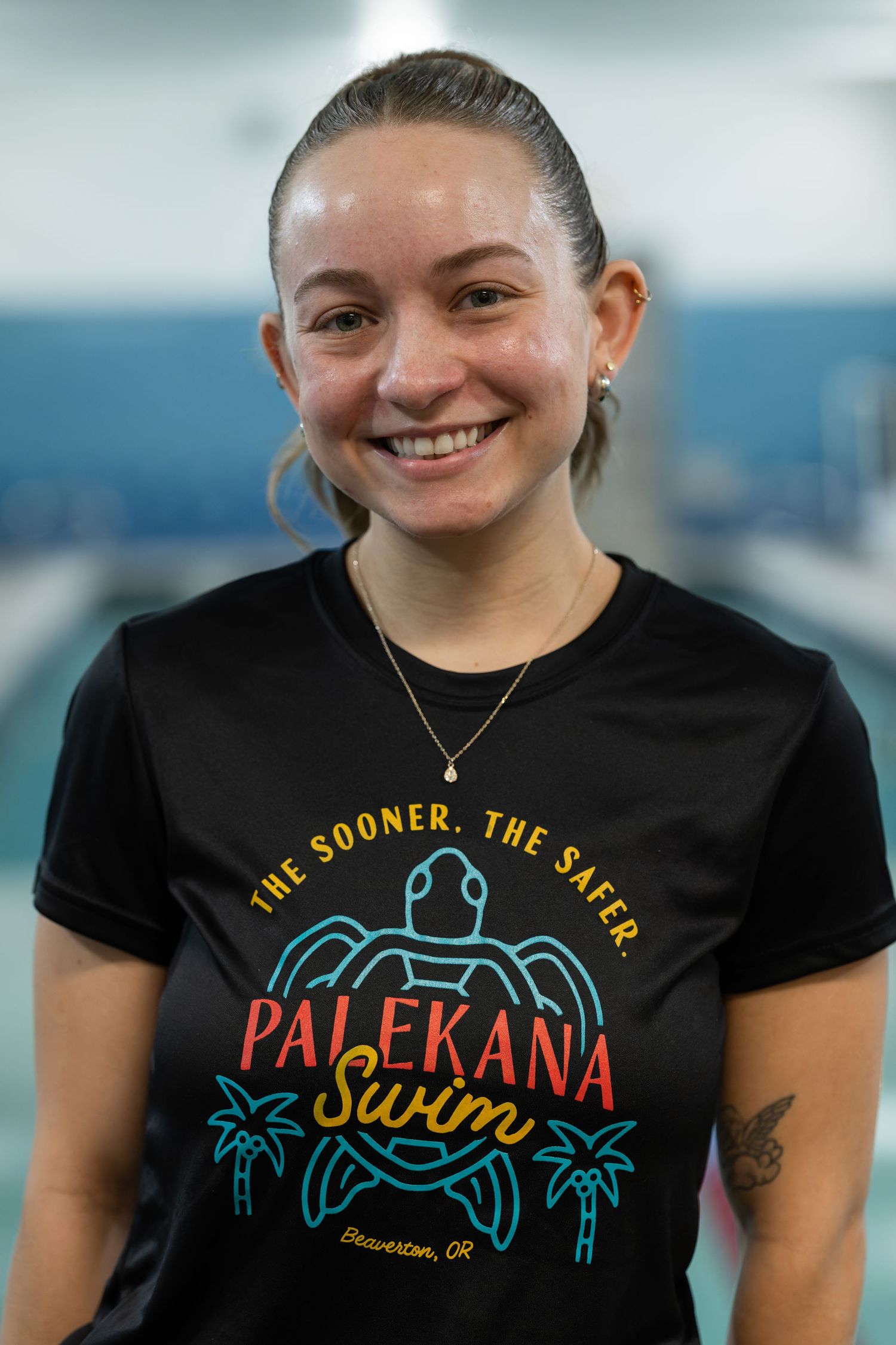 Smiling woman wearing a black Palekana Swim t-shirt standing near an indoor pool.