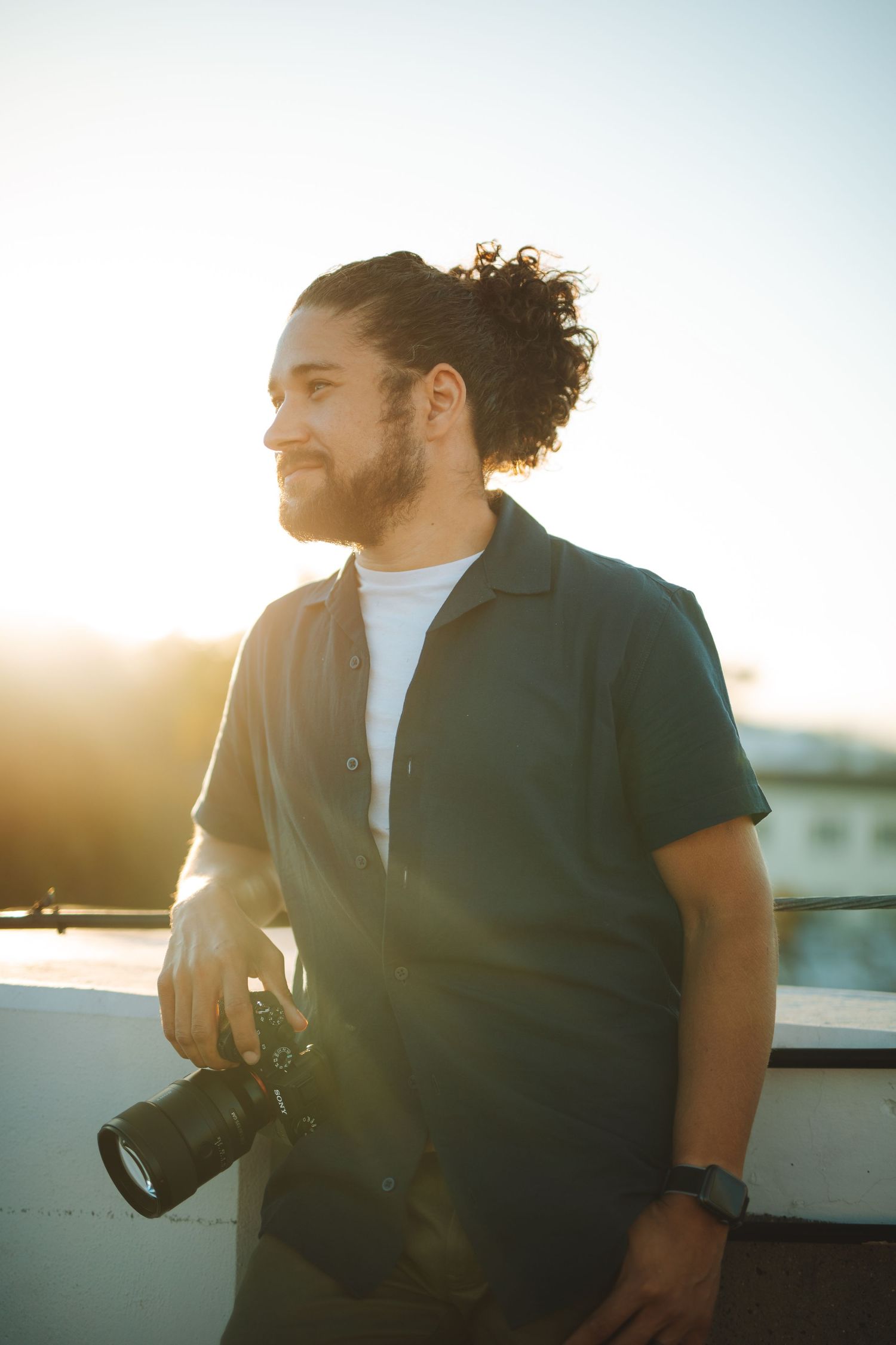 Bearded man holding camera looking sideways in bright sunlight wearing a dark shirt outdoors at sunset.