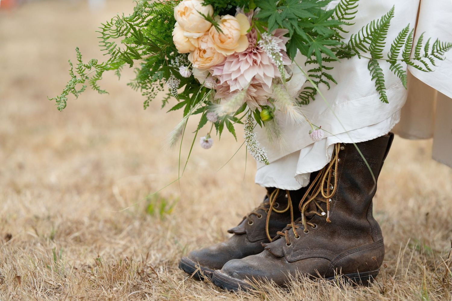 Rustic bridal boots with lace-up details paired with a lush bouquet of pink roses and ferns on dry grass.