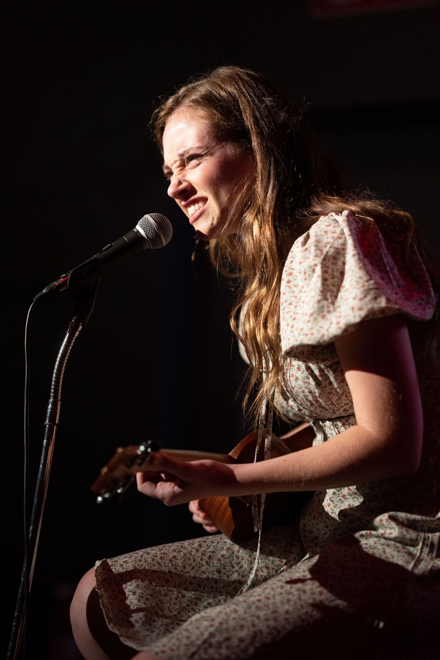 Young woman making a grimacing face and playing ukulele while singing into microphone on stage in floral dress at comedy show