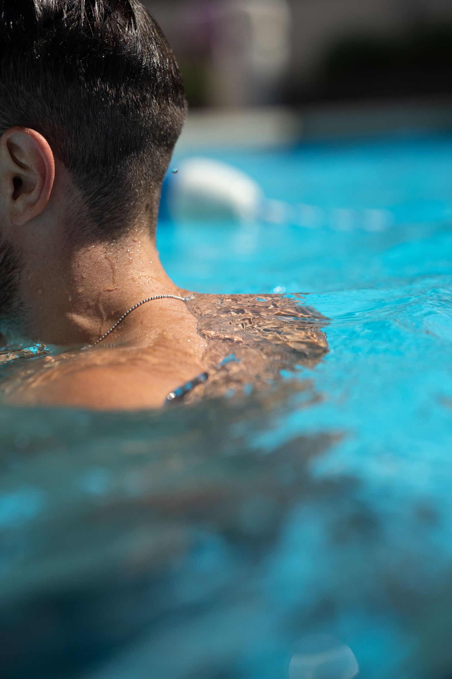 Close-up rear view of a man with a necklace relaxing in a turquoise swimming pool.