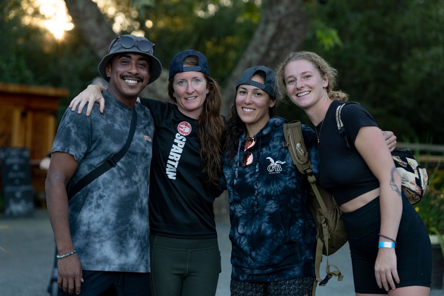 Four diverse friends smiling and posing together outdoors at an evening event, wearing casual black and grey tie-dye and athletic clothing.
