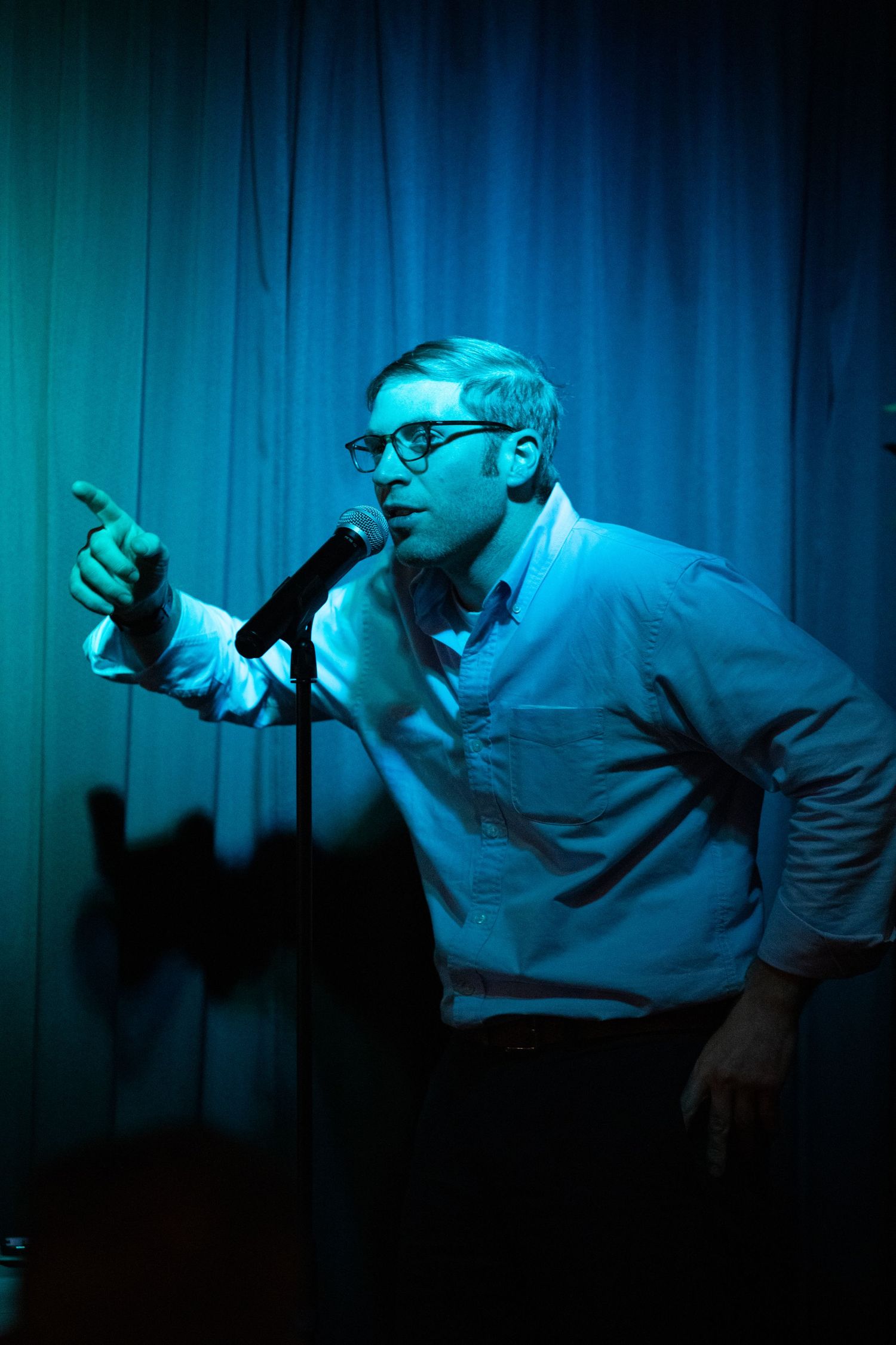 Comedian Joe Pera performing stand-up comedy at microphone under blue stage lighting on stage at an event in San Luis Obispo, California