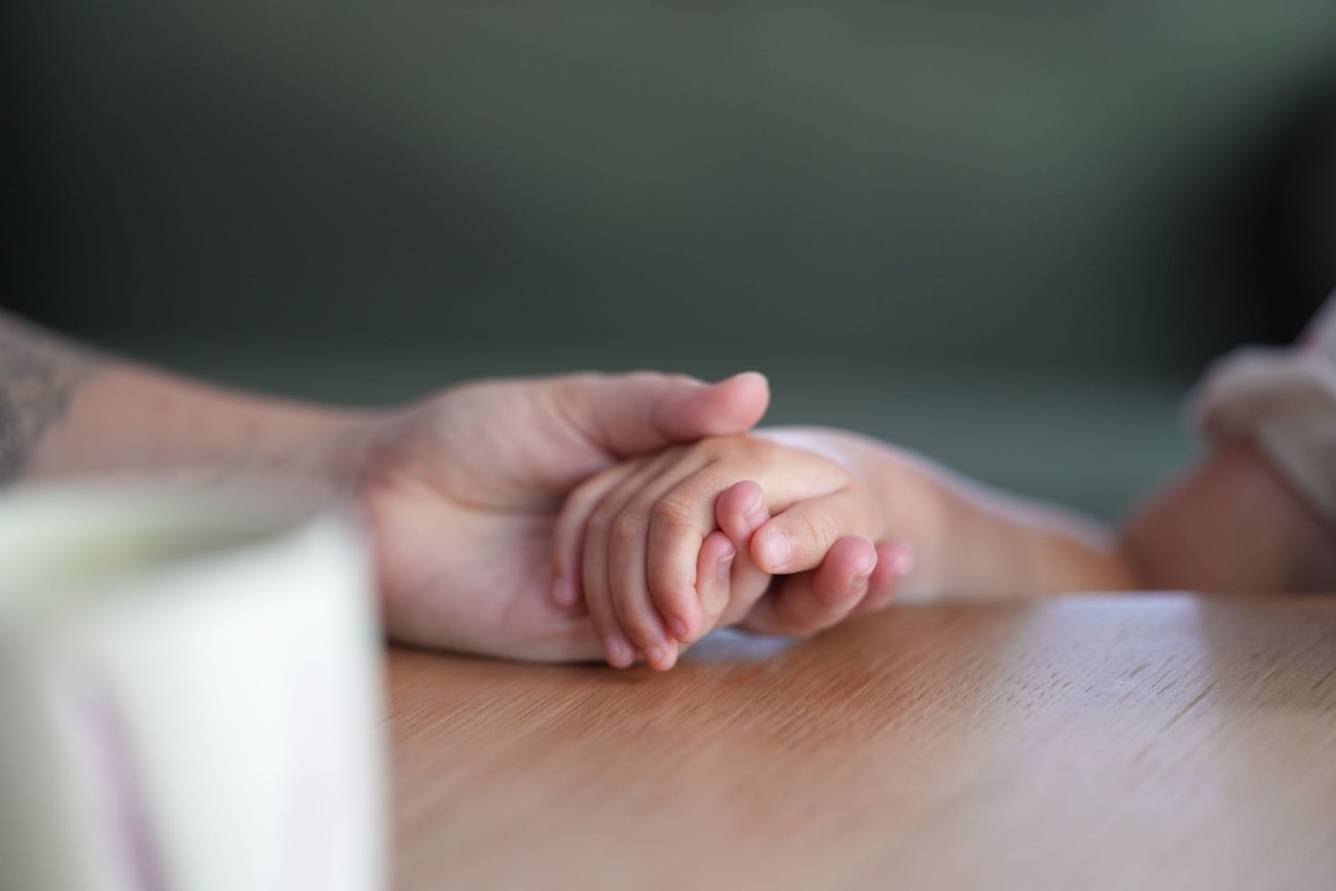 Adult hand gently holding a small baby hand on a wooden table surface.