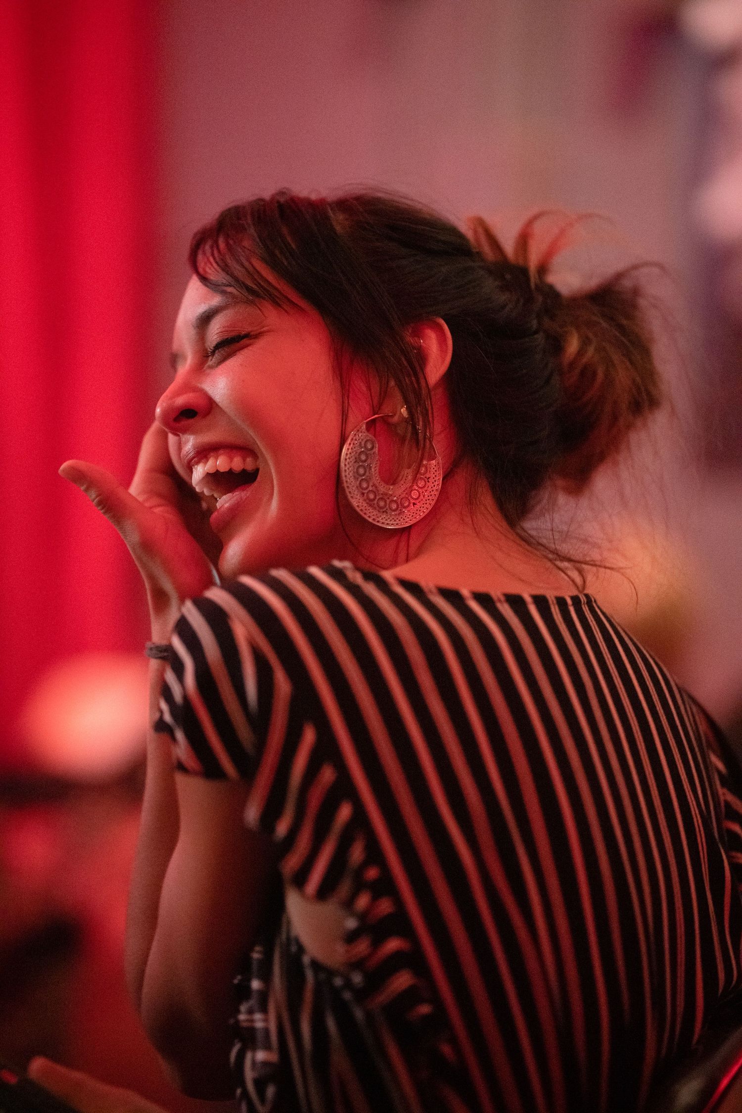 Laughing woman with large hoop earrings in striped top enjoying a comedy show under red lights.