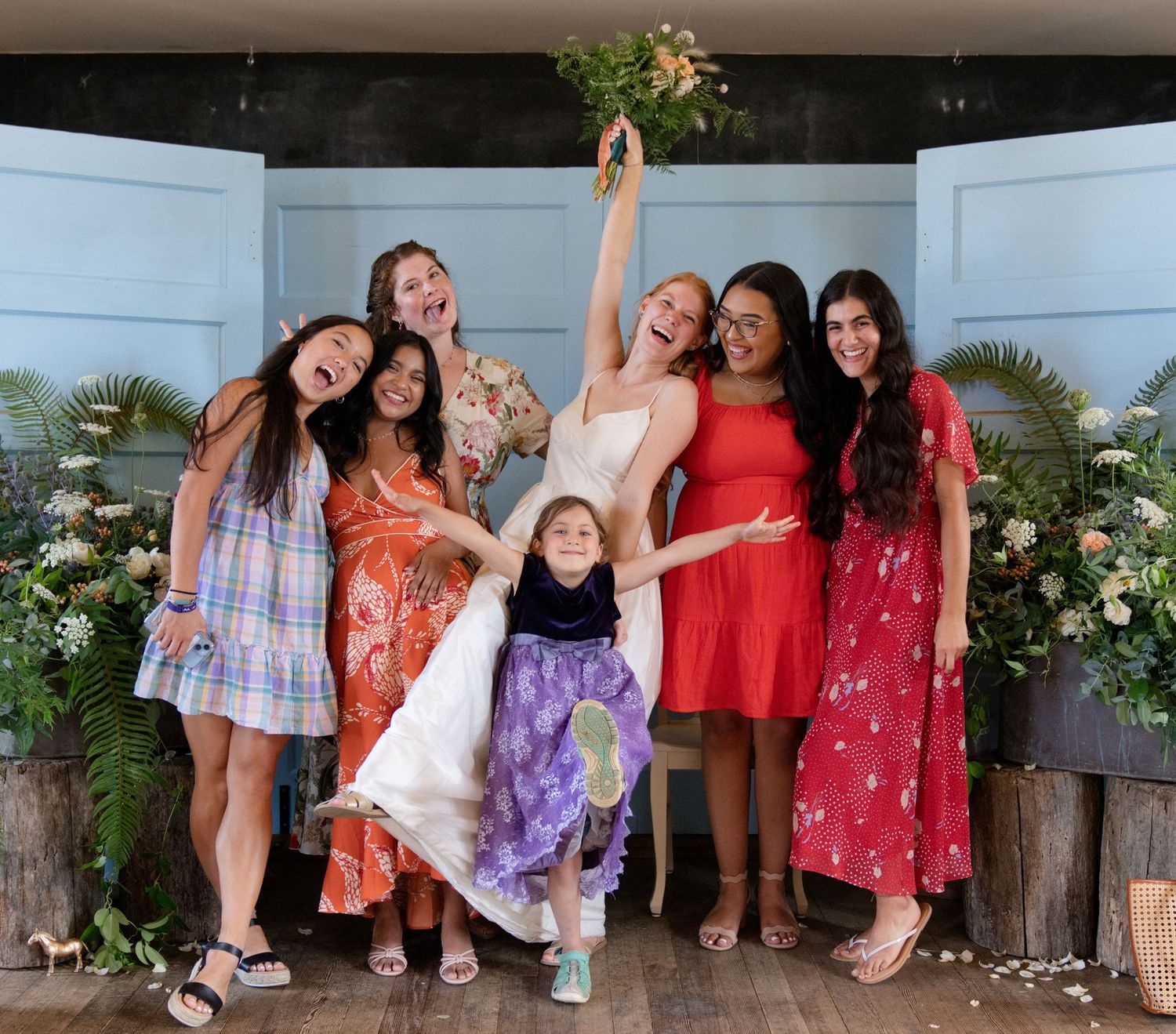 Happy bride holding bouquet aloft surrounded by smiling bridesmaids in colorful dresses indoors.