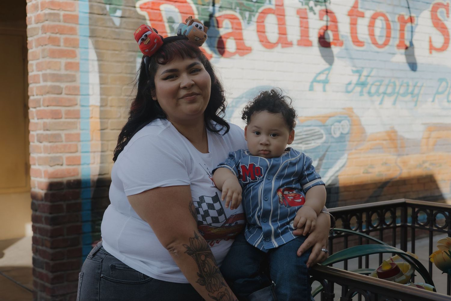 A woman with Mickey Mouse ears holds a baby boy near a colorful Radiator Springs mural at Disneyland.