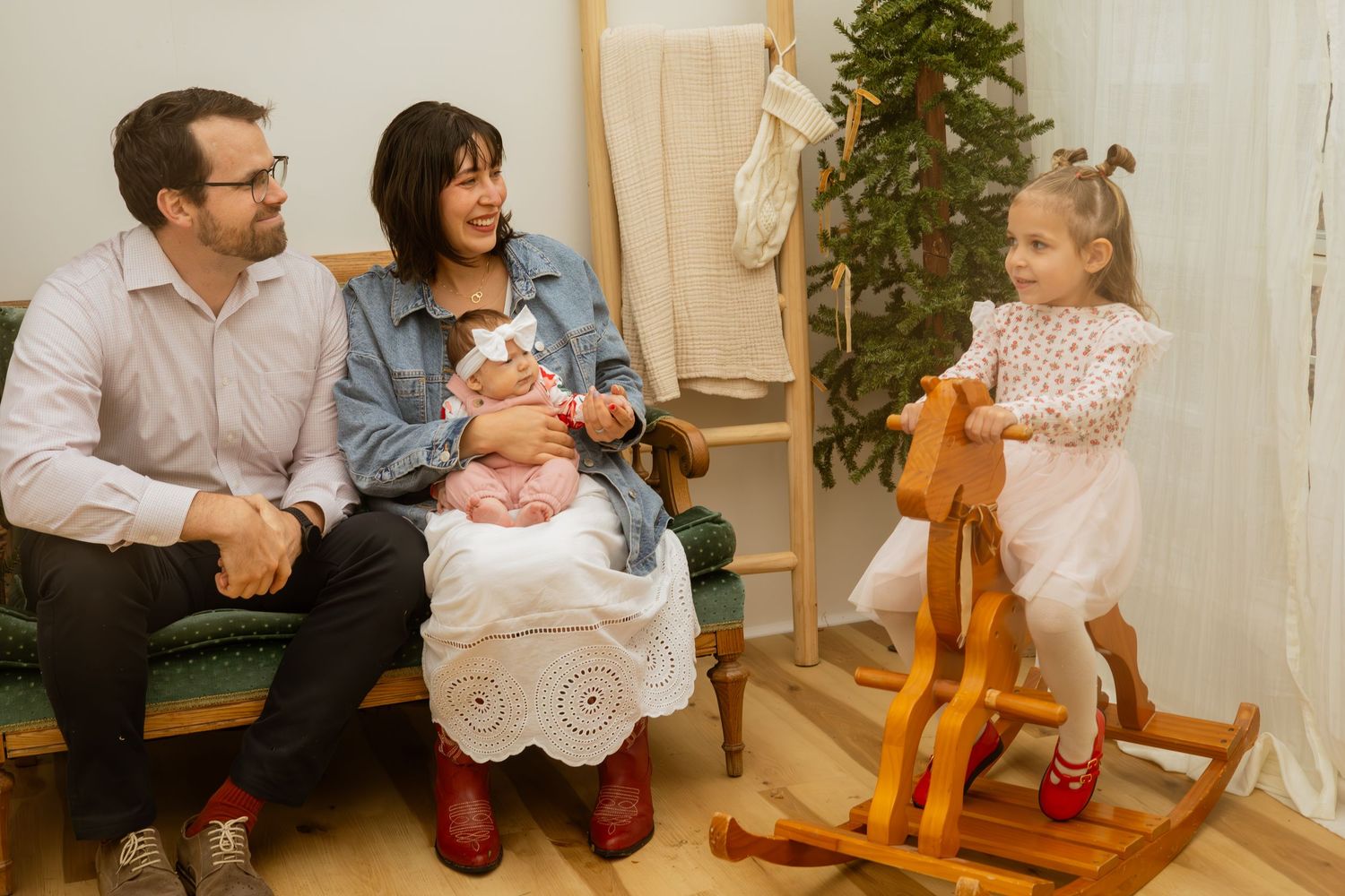 Family with newborn baby and toddler girl on rocking horse in cozy Christmas decorated room.