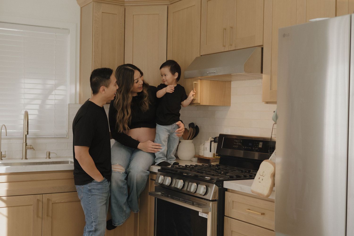 Family of three sharing a warm moment in a modern kitchen with light wood cabinets and stainless steel appliances.