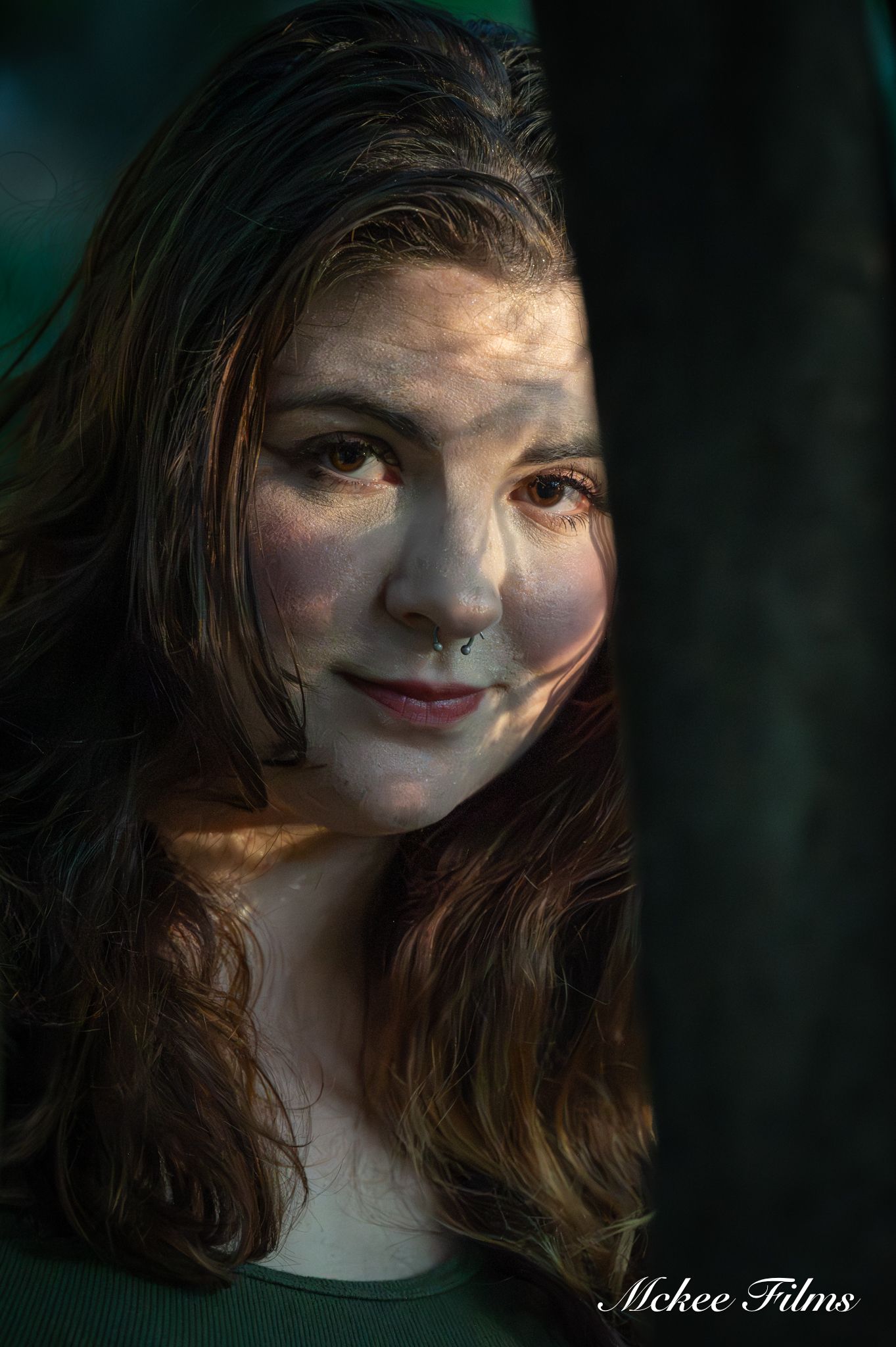 Young woman with long brown hair and nose ring smiling softly in dappled sunlight among trees, portrait by Mckee Films.