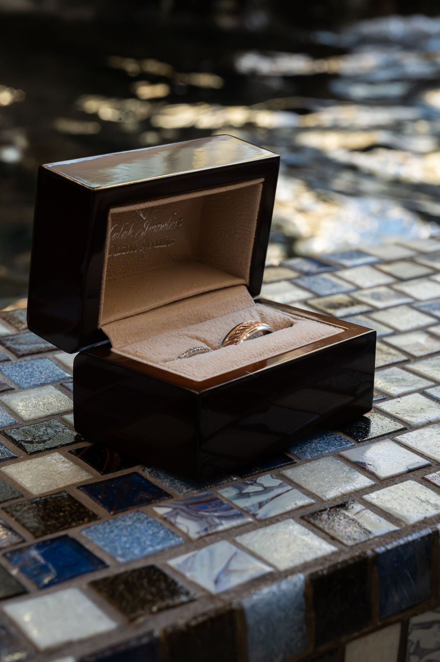 A gold wedding ring displayed in an open dark wood jewelry box on a mosaic tile surface.