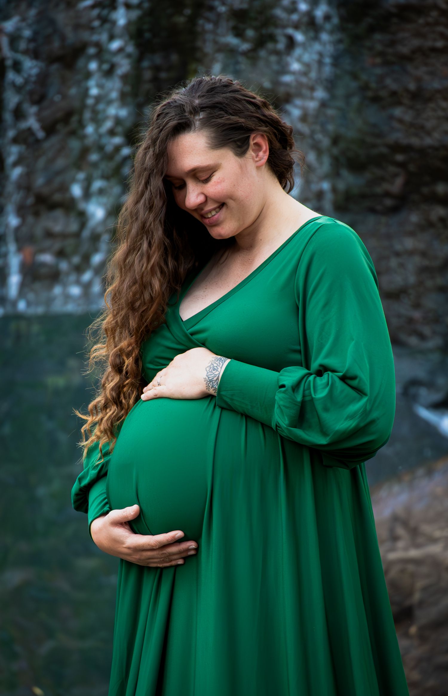 Smiling pregnant woman in green dress cradling baby bump outdoors near waterfall with long curly brown hair.